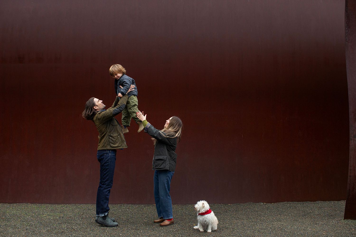 Family with child on shoulders and small white dog pose outdoors against a large red metal wall.