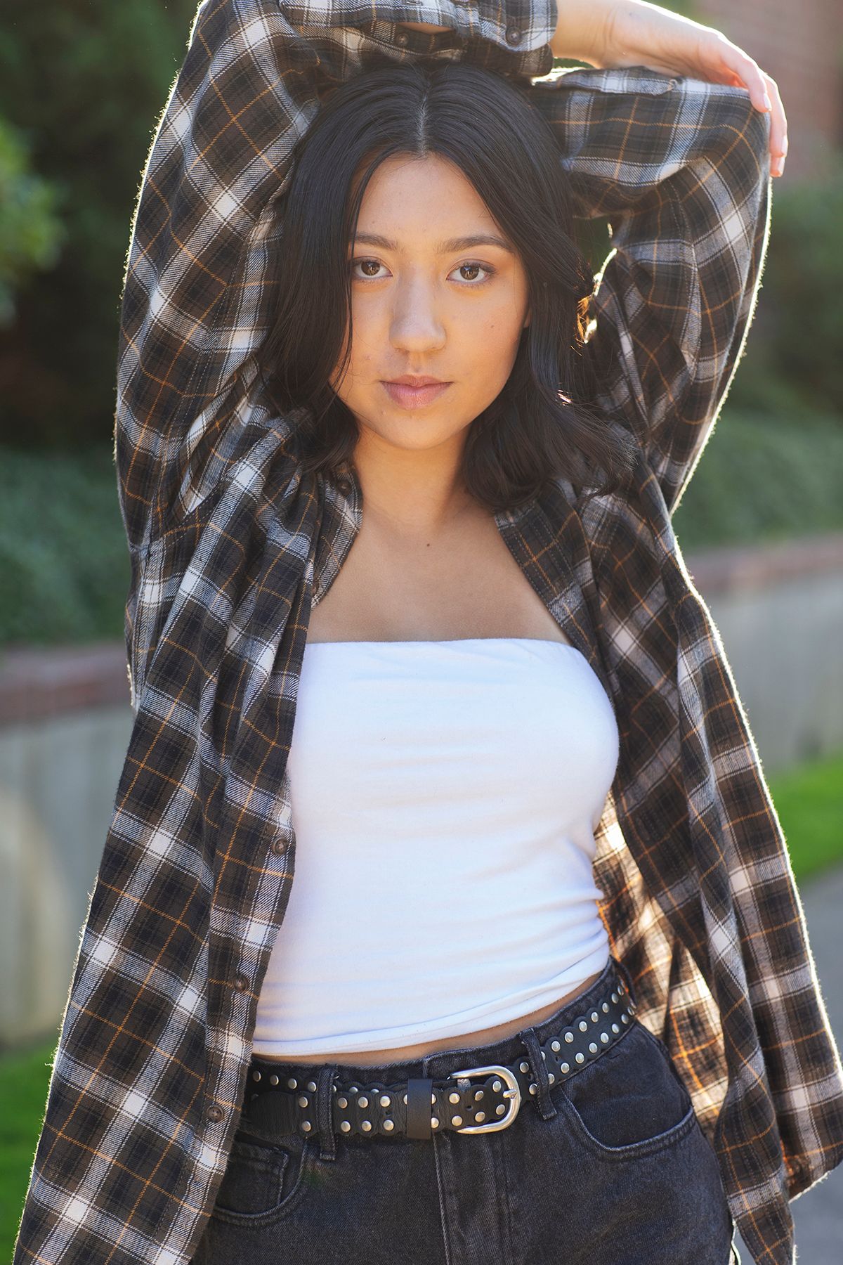 Young woman in white tube top, plaid shirt, and jeans poses outdoors with arms raised, sunlight highlighting her dark hair.