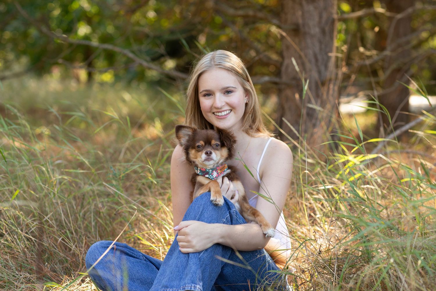 Smiling young woman sitting in tall grass holding small Chihuahua dog outdoors in nature.