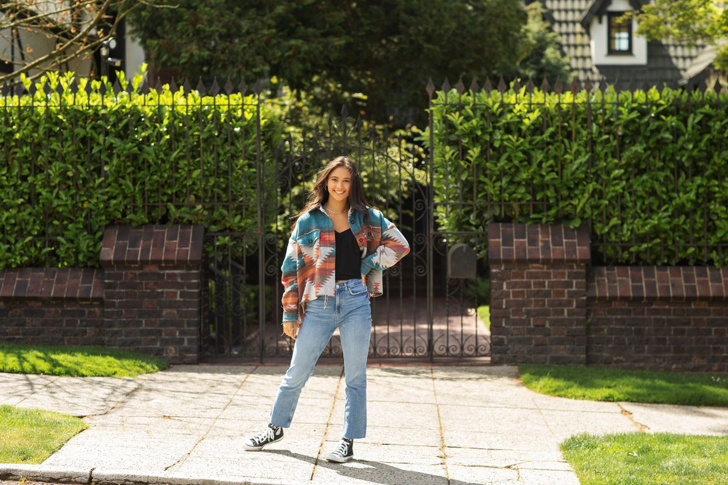 Young woman in colorful patterned jacket posing confidently by an iron gate on a sunny day.