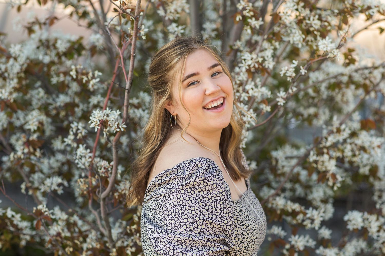 Young woman smiling joyfully outdoors among white spring blossoms, wearing a floral off-shoulder top.