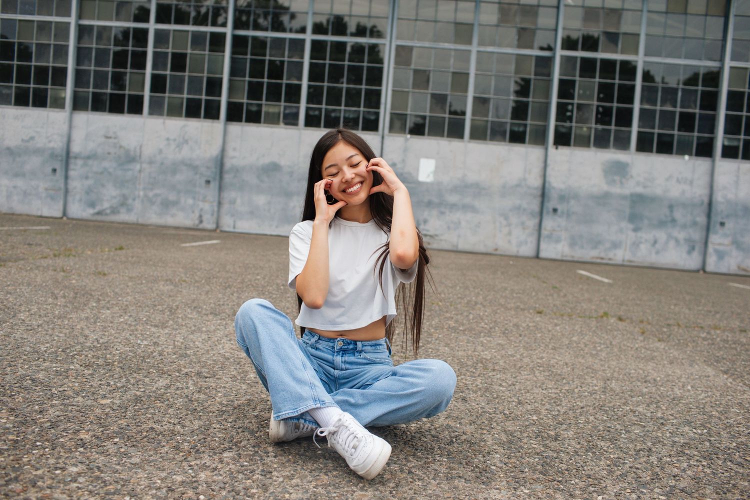 Smiling young woman sitting cross-legged on pavement making peace signs in white top and jeans.