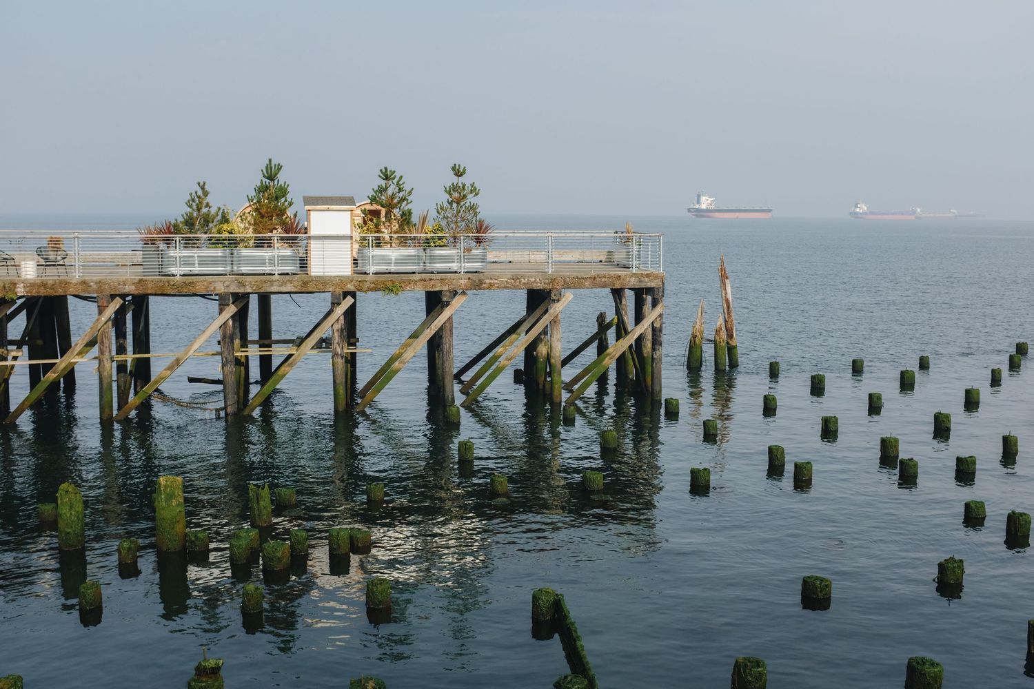 A weathered wooden pier extends over calm water with old pilings reflected below on a hazy day.
