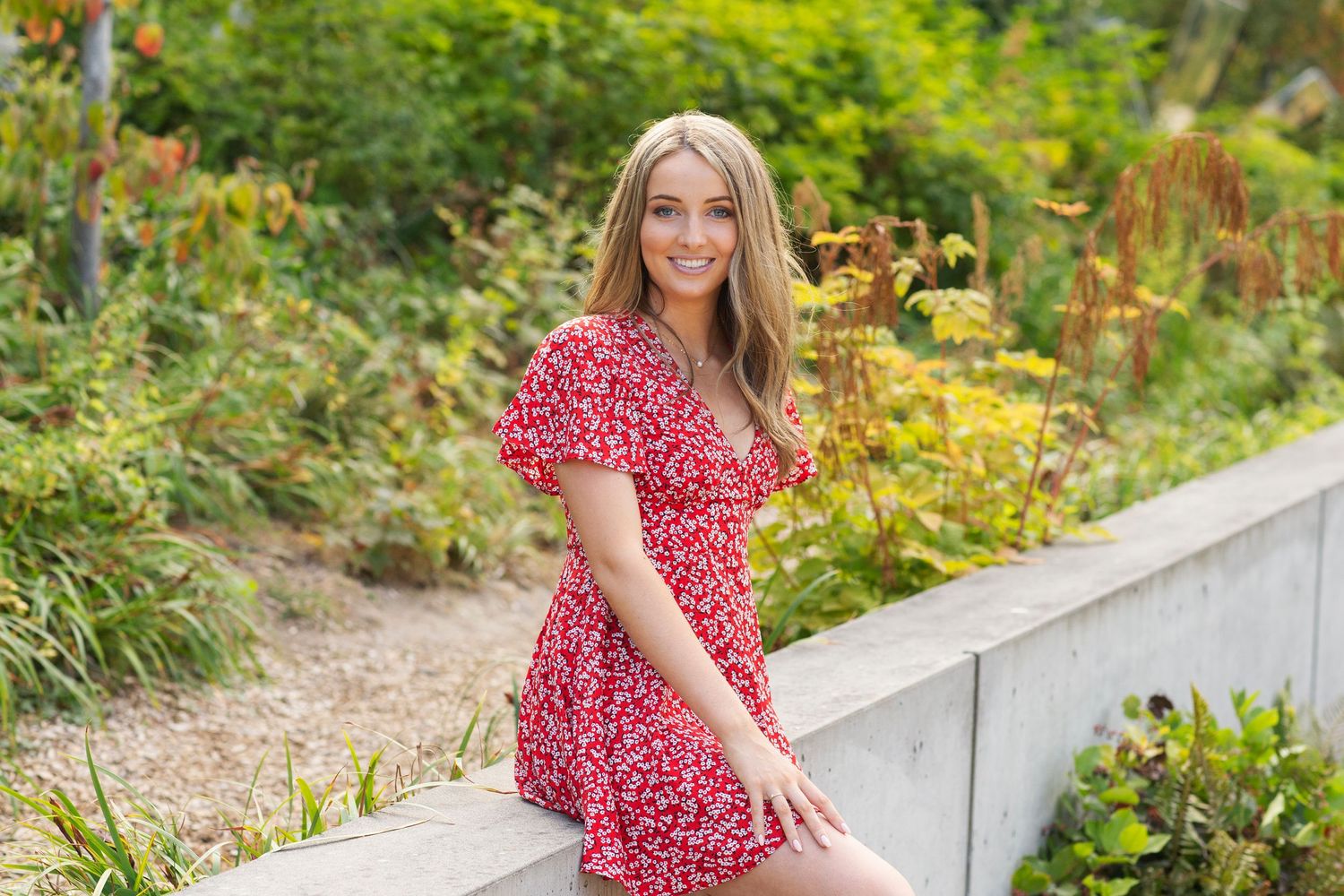 Smiling blonde woman in red floral dress sitting on a garden wall outdoors.