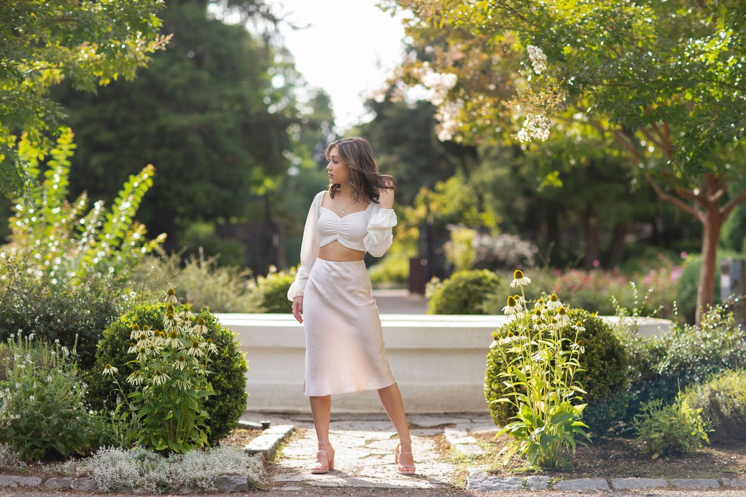 Young woman in a white crop top and blush midi skirt posing in a lush garden with stone fountain.