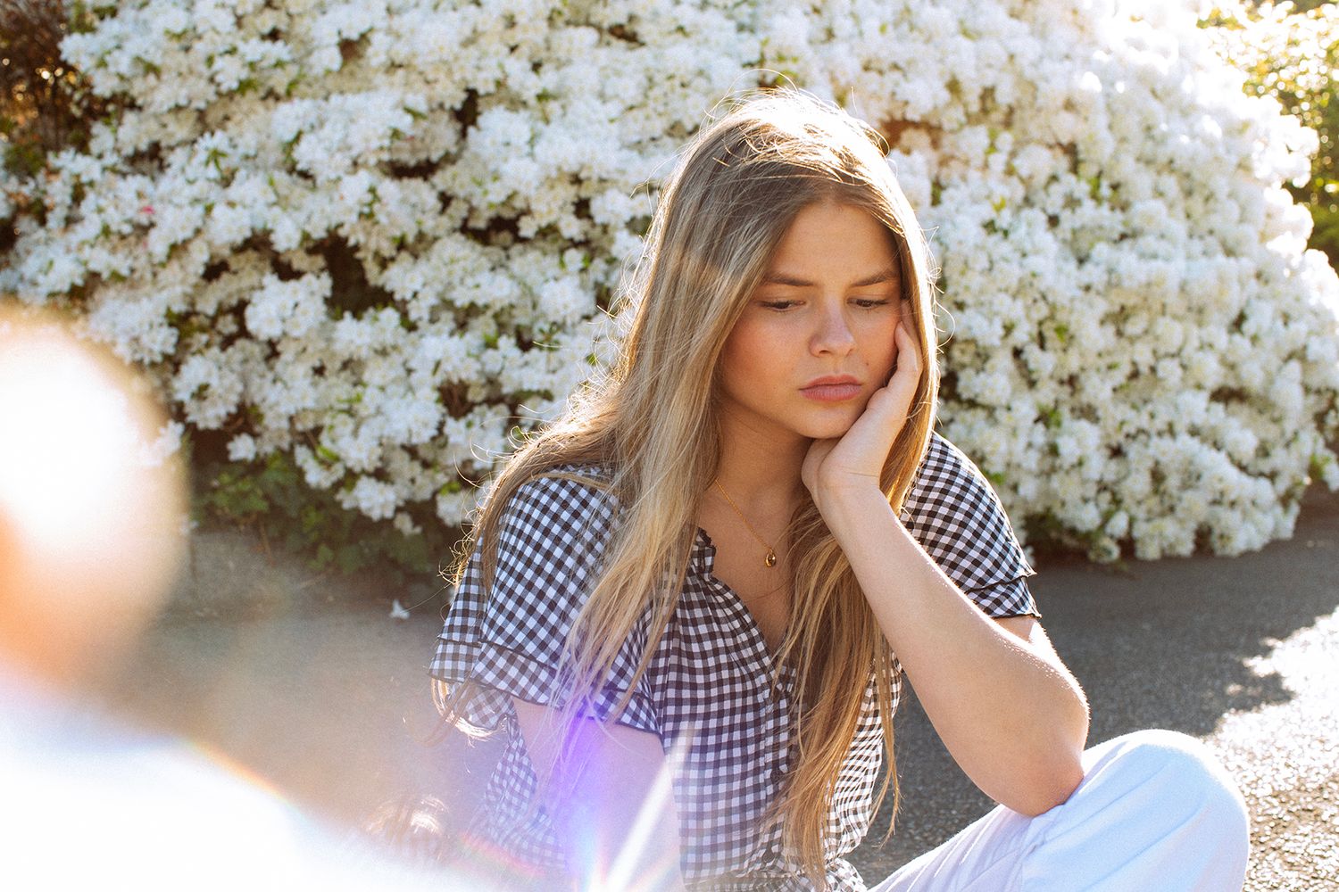 Young woman in gingham shirt sitting outdoors near white flowering bushes, resting her cheek on her hand.