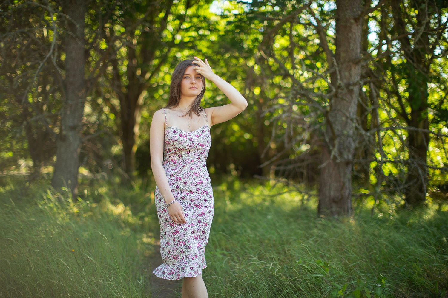 Young woman in floral sundress standing in lush green forest, shielding eyes from sunlight on a bright summer day.