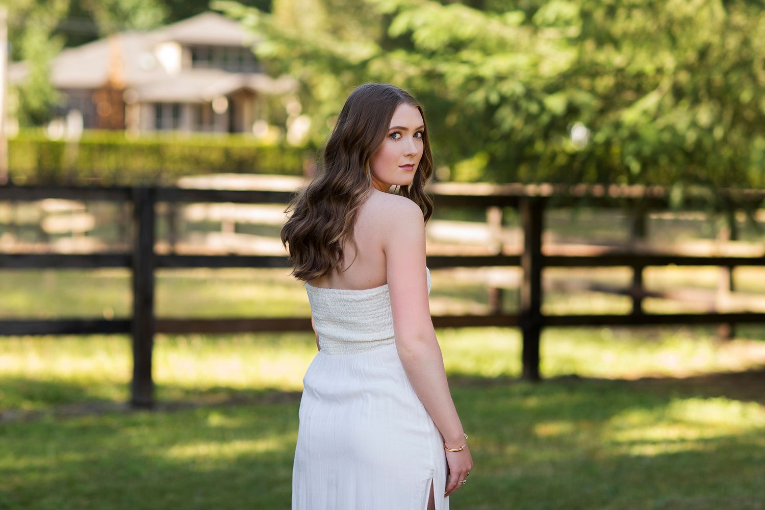 Young woman in white strapless dress posing outdoors near wooden fence with lush green trees in background.