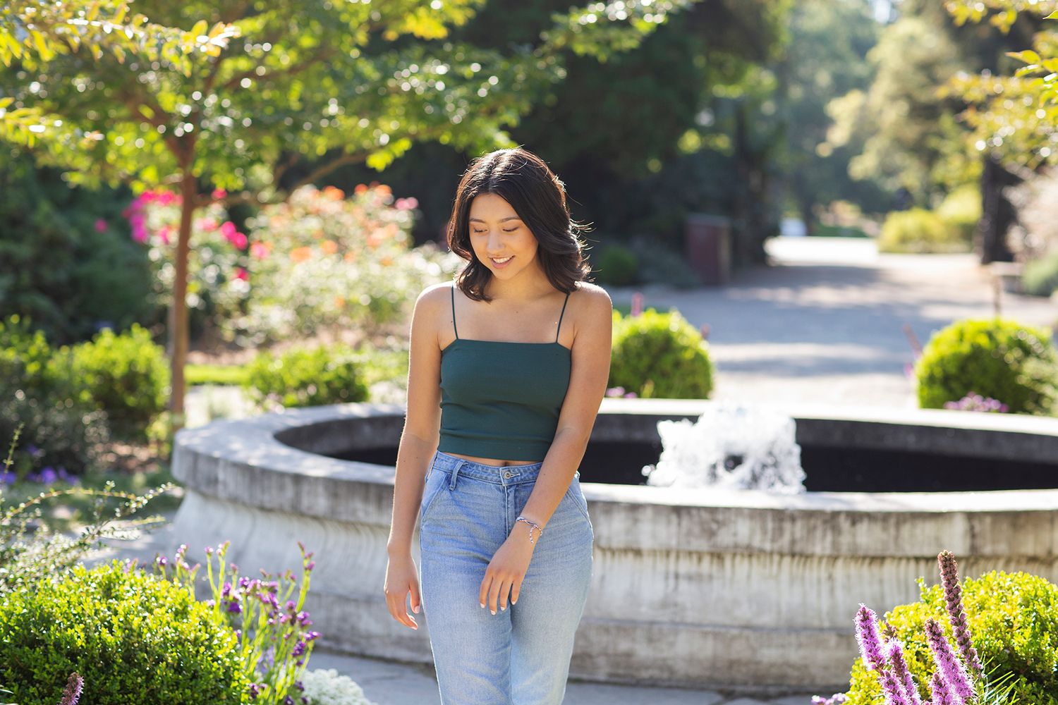 Young woman in green crop top and jeans smiling in a sunny garden with a fountain backdrop.