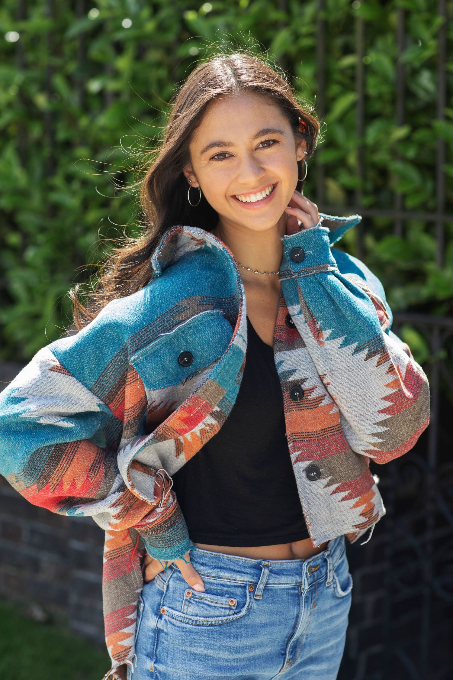 Smiling young woman wearing a colorful aztec print jacket with jeans in a sunny garden.