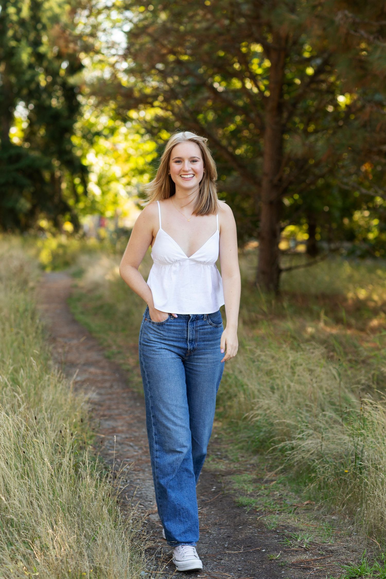 Smiling blonde woman walking on a dirt path in white cami top and jeans surrounded by green trees.
