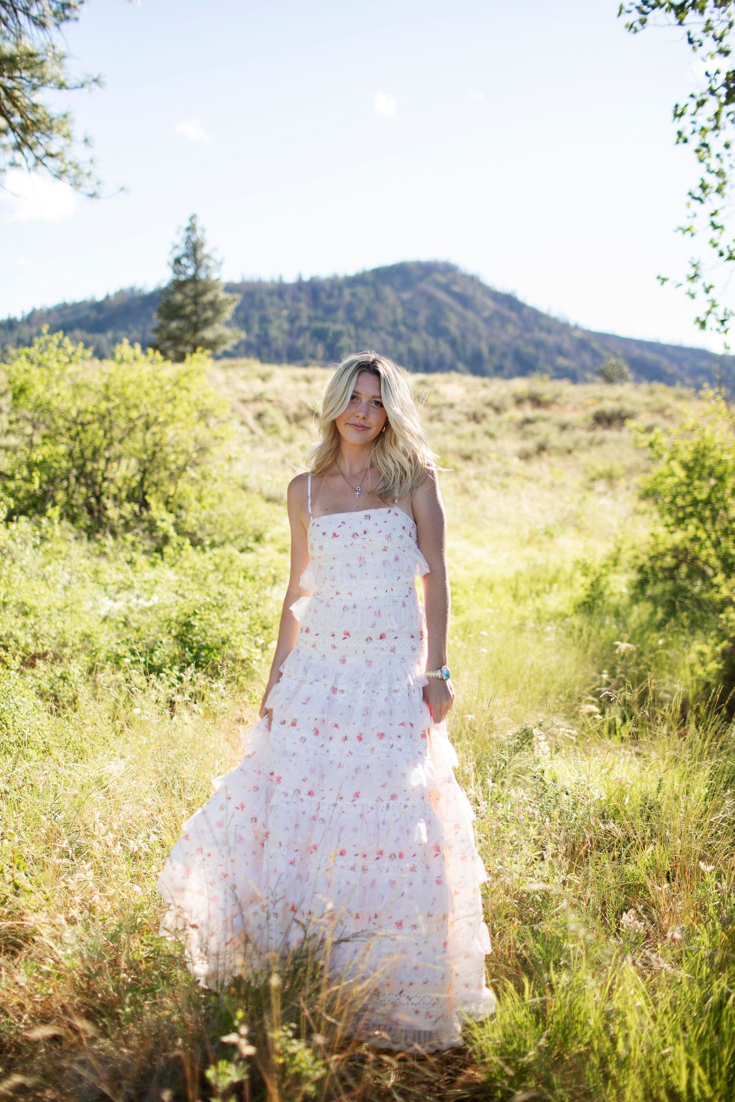 Blonde woman in floral tiered maxi dress standing in a green meadow with mountain backdrop.