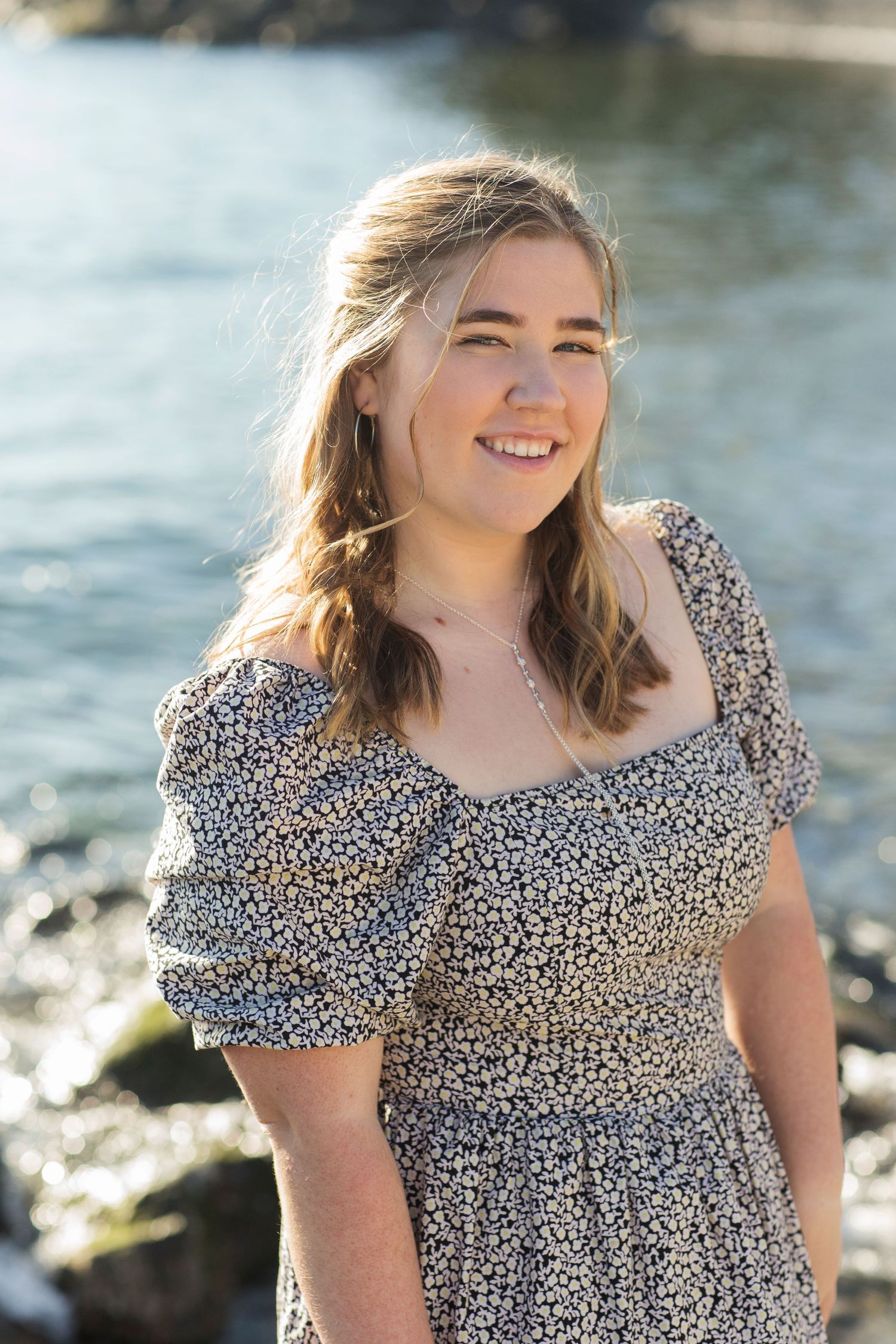 Young woman in floral dress smiling outdoors near rocky shoreline with sunlit water in background.