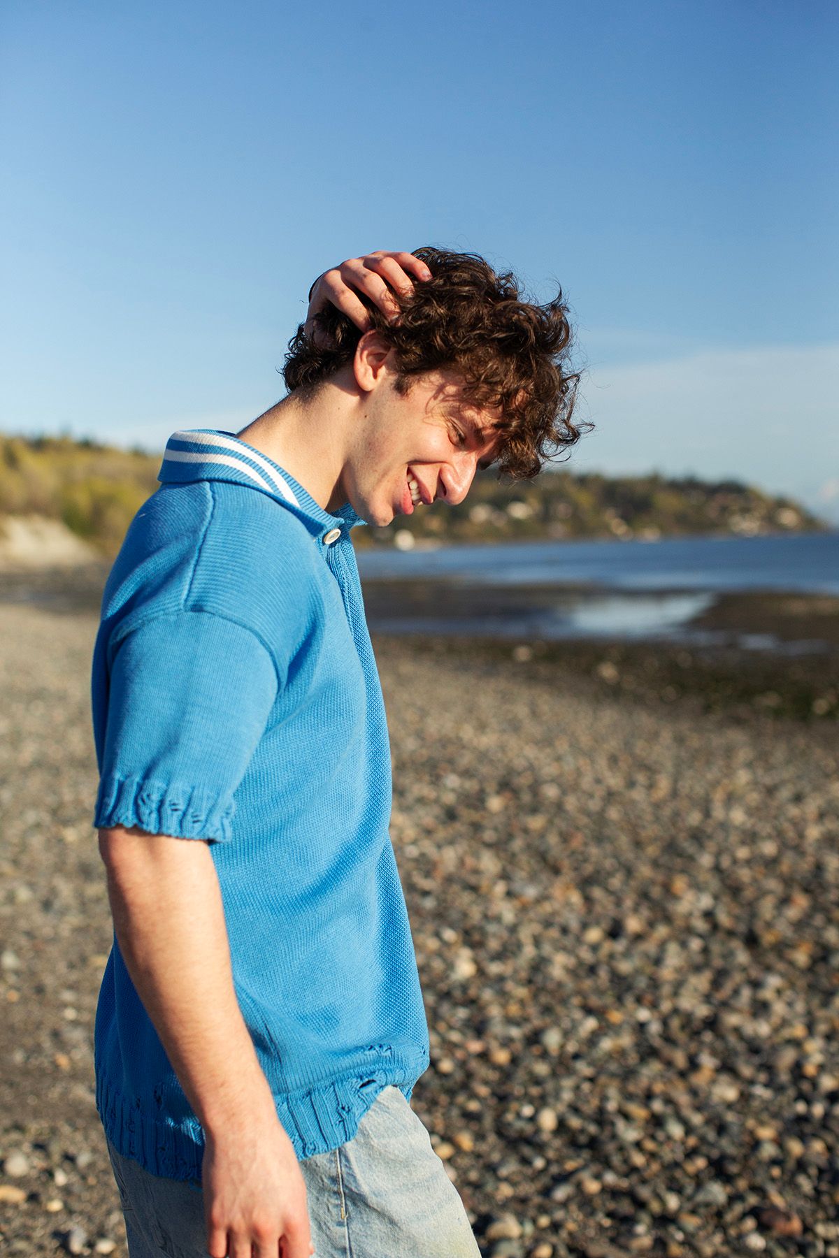 Young man with curly hair laughing on a rocky beach, wearing a blue polo shirt on a sunny day.