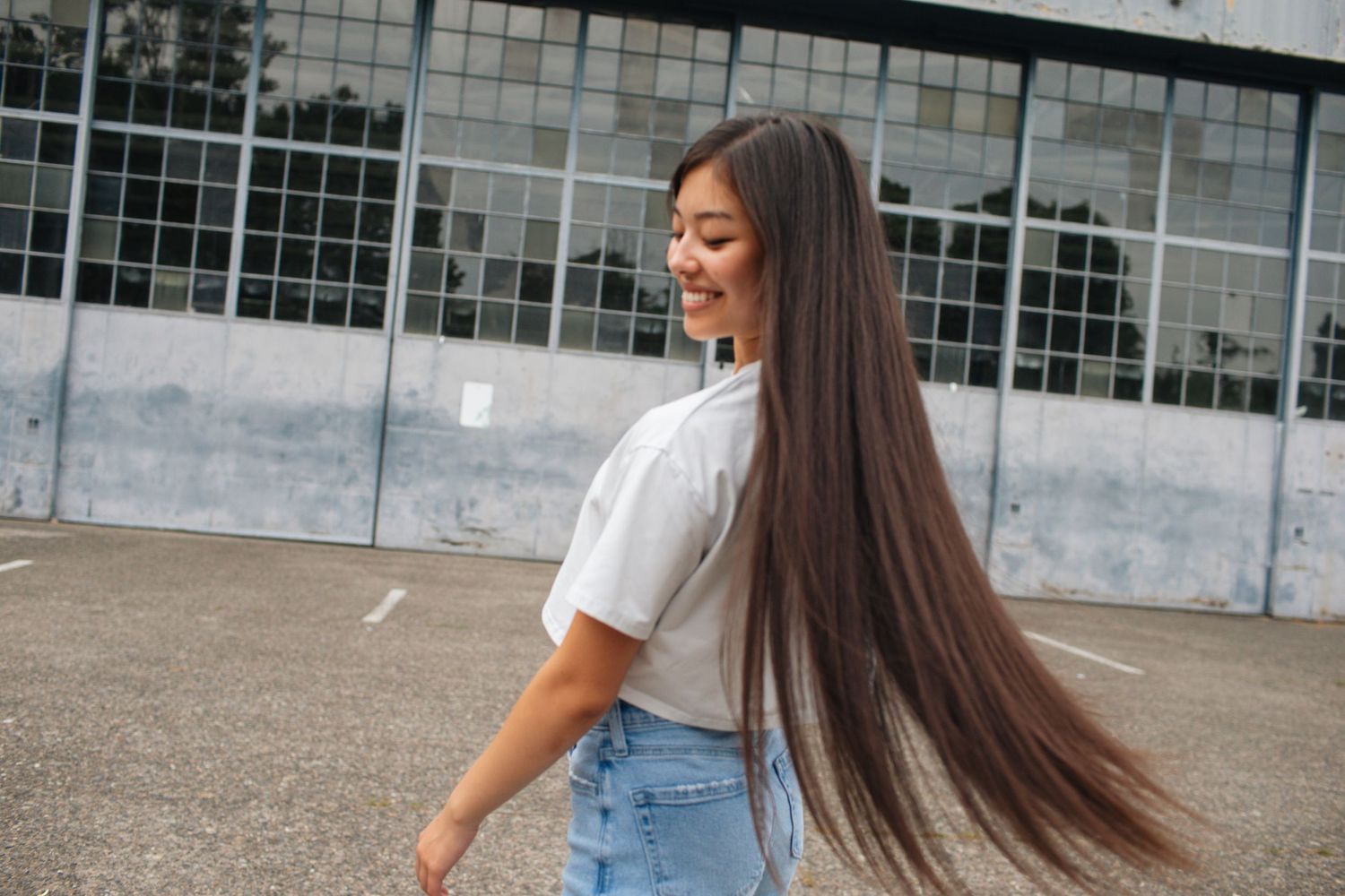Young woman with long flowing dark hair smiling in white top and jeans in urban outdoor location.
