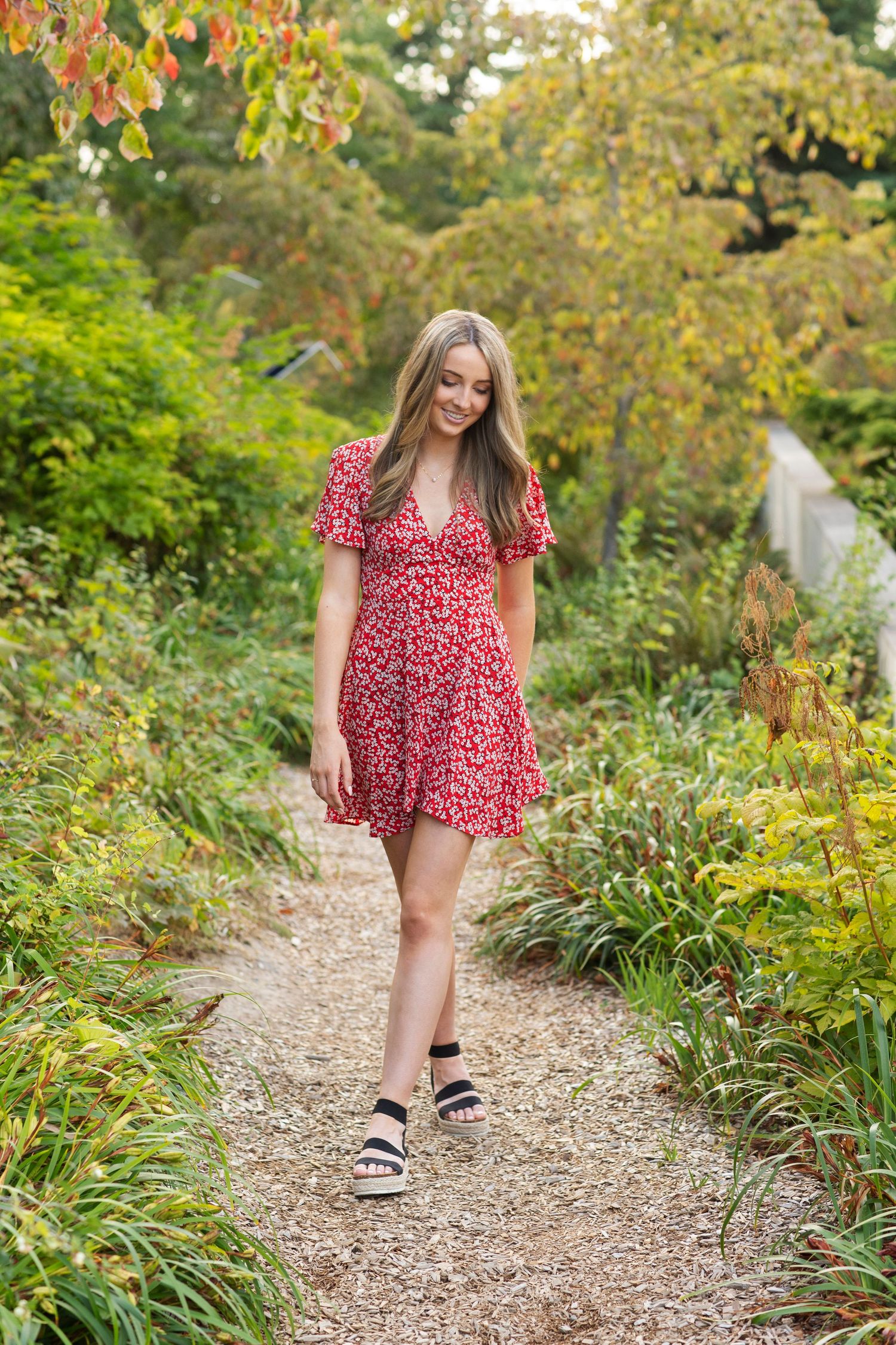 Young woman in red floral dress walking along a lush garden path in autumn.