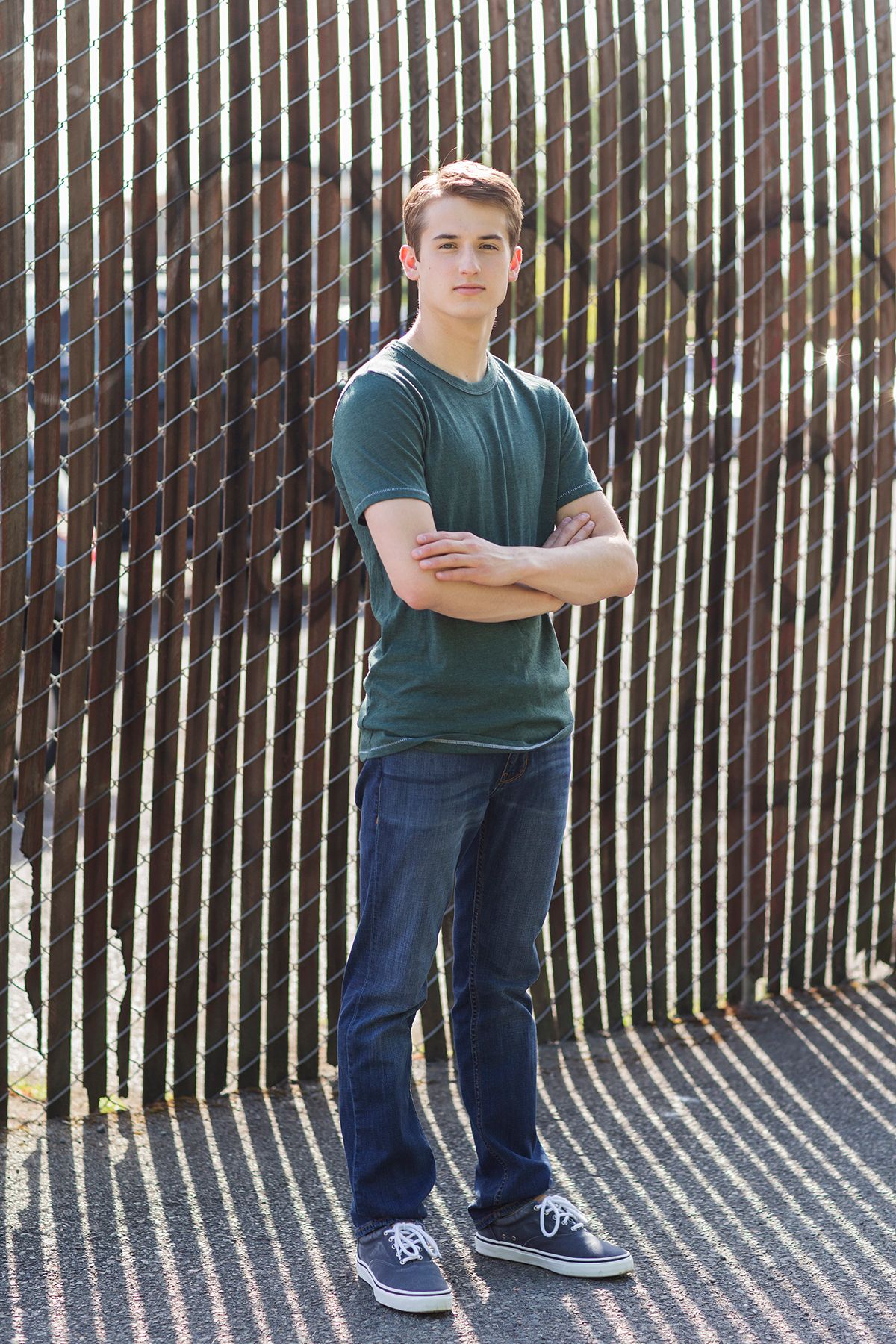 Teen boy in green t-shirt and jeans stands with arms crossed against a diagonal fence outdoors.