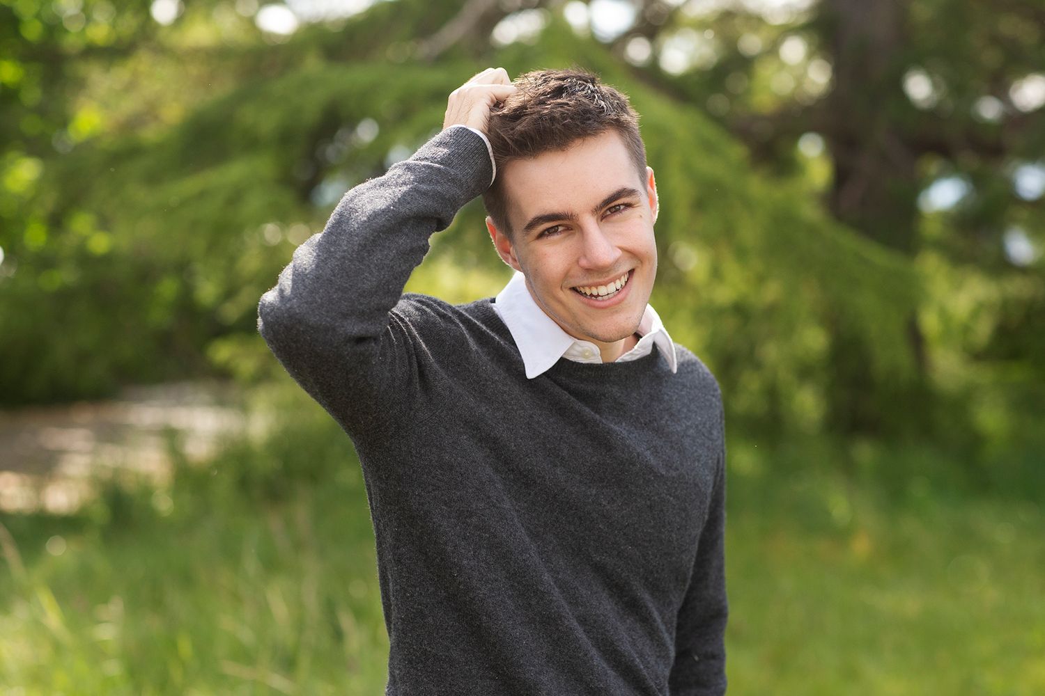 Smiling young man in gray sweater posing outdoors with hand on head against lush green trees.