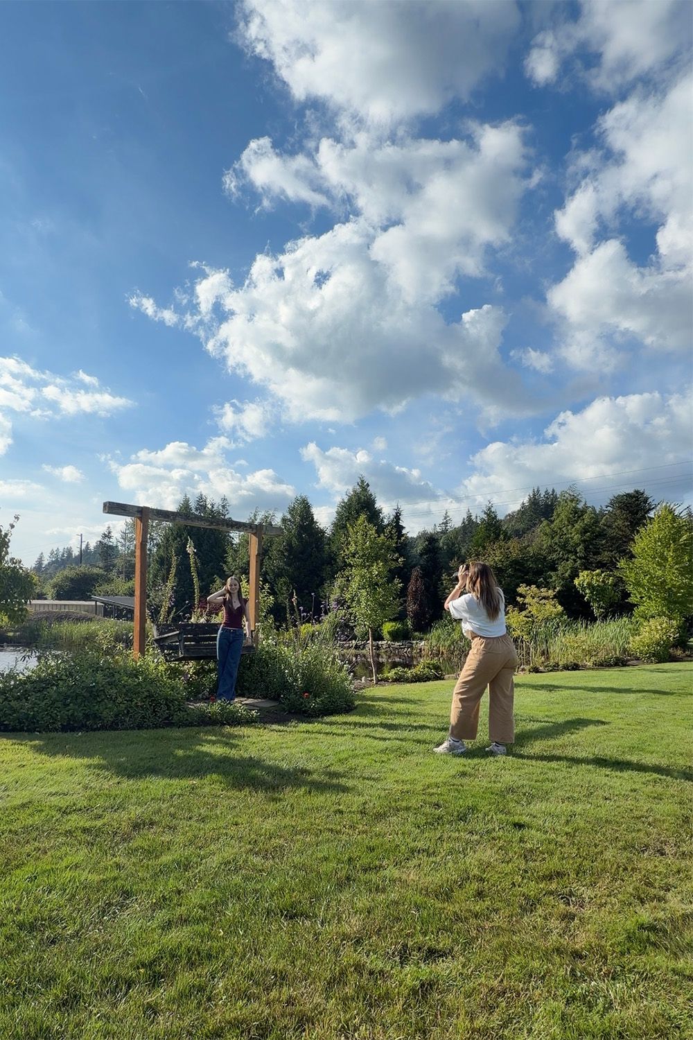 Two women in a lush garden, one photographing the other near a wooden arch under a blue sky with fluffy clouds.