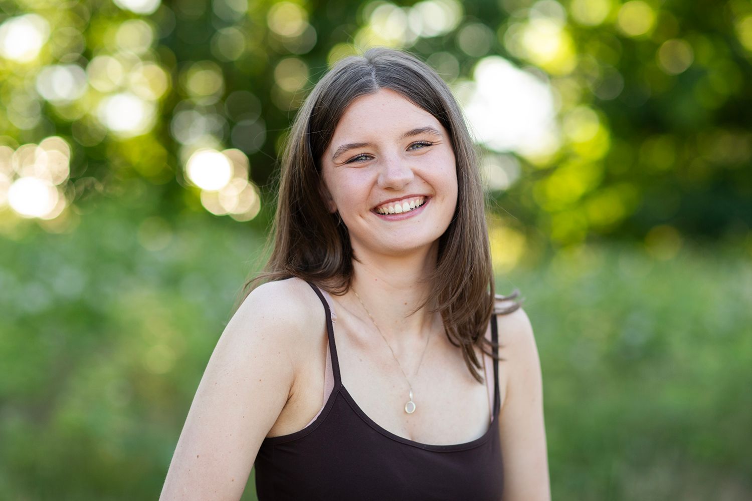 Smiling young woman with brown hair wearing a black tank top and gold necklace outdoors with green trees.