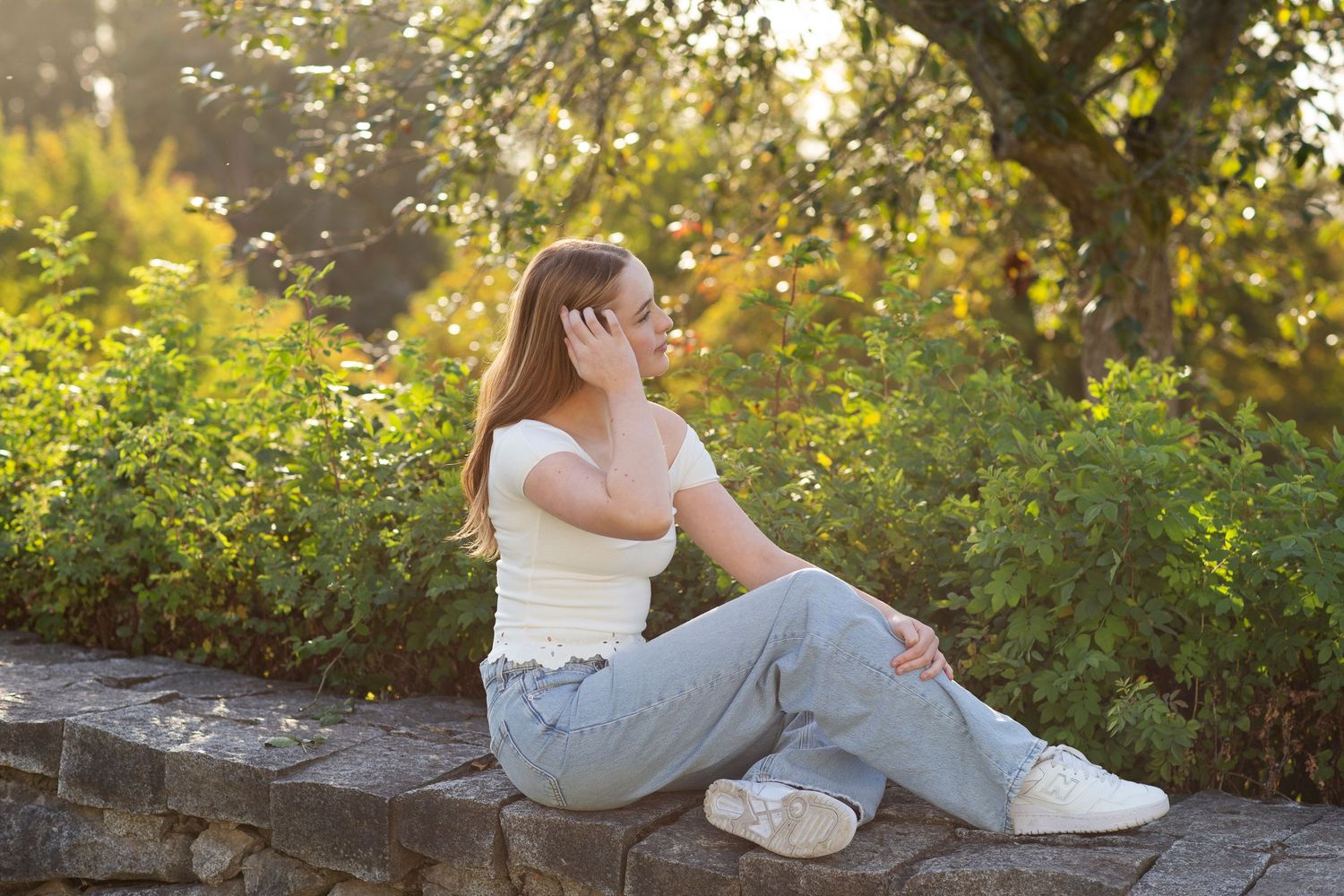 Young woman in white top and jeans sitting on stone wall surrounded by lush green bushes in golden sunlight.
