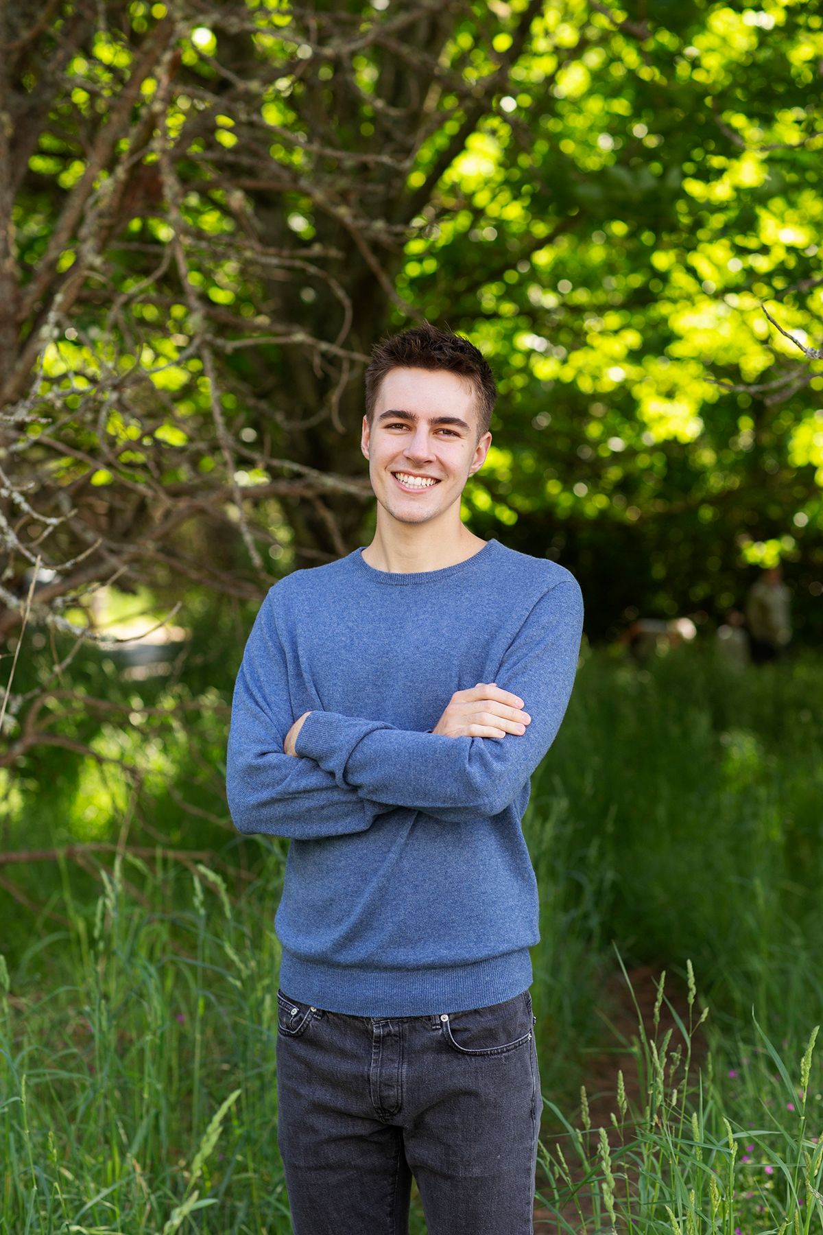 Young man in blue sweater smiling with arms crossed outdoors among lush green trees and tall grass.