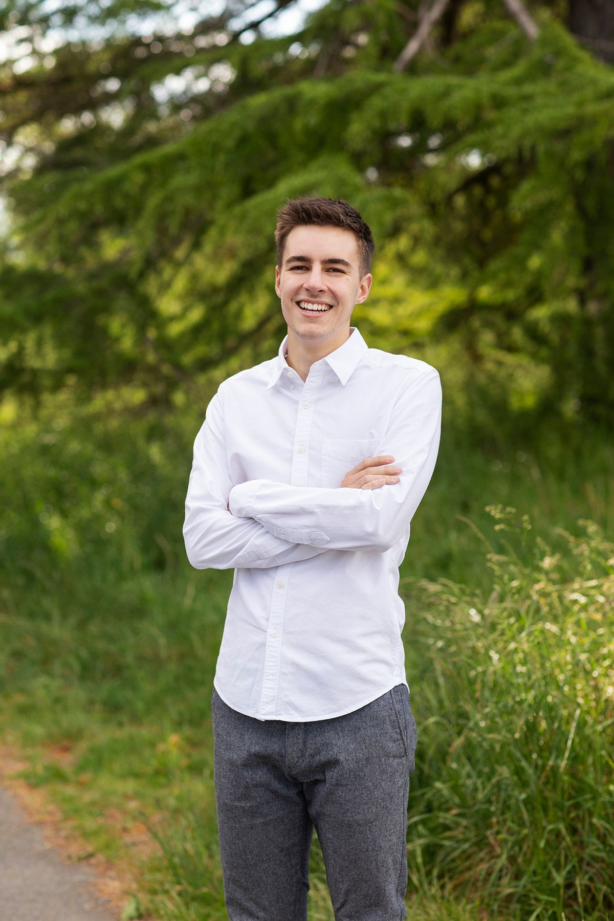 Young man in white shirt and gray pants stands outdoors with arms crossed, smiling confidently among green trees.