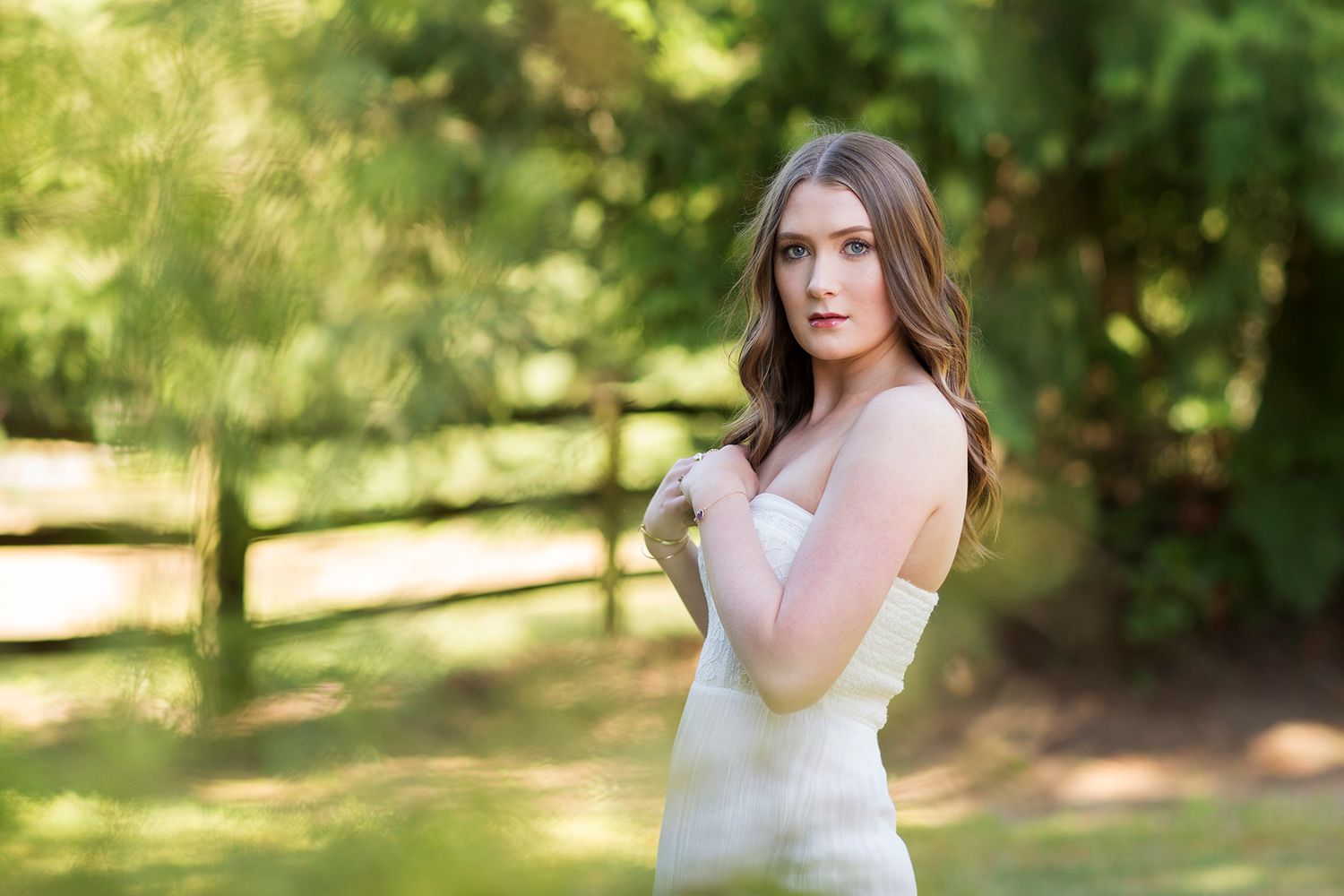 Young woman in white strapless dress posing outdoors with lush green trees and wooden fence in background.