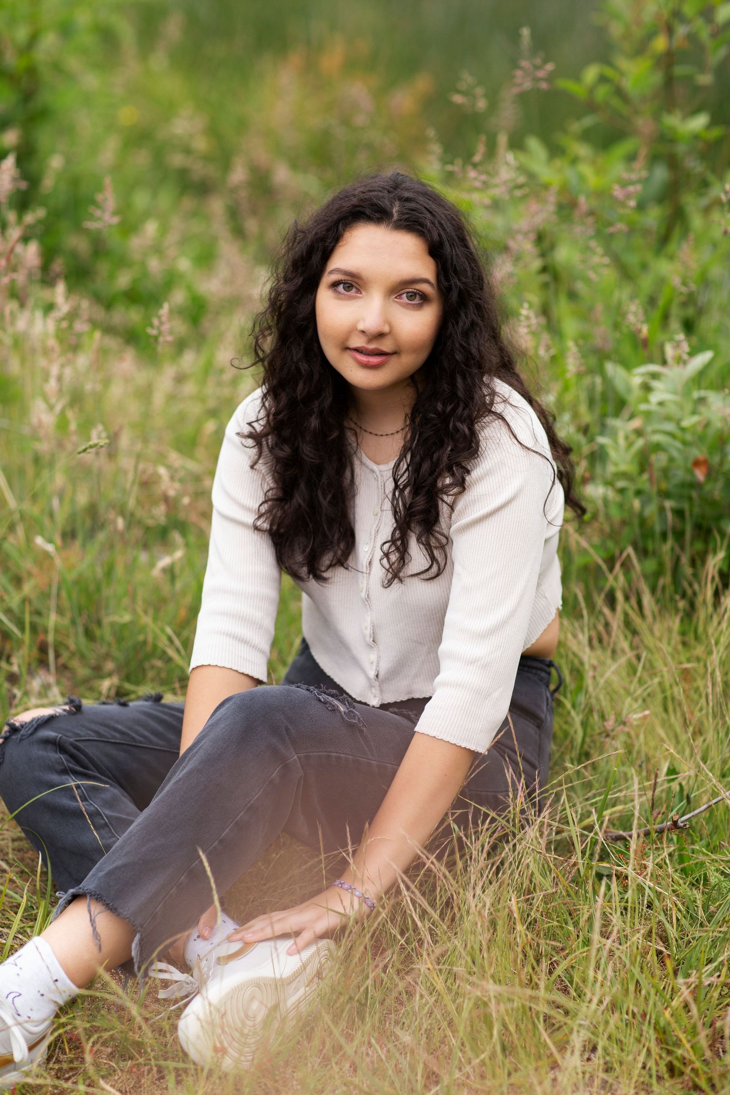 Young woman in white cardigan sitting in a grassy field with curly dark hair smiling.