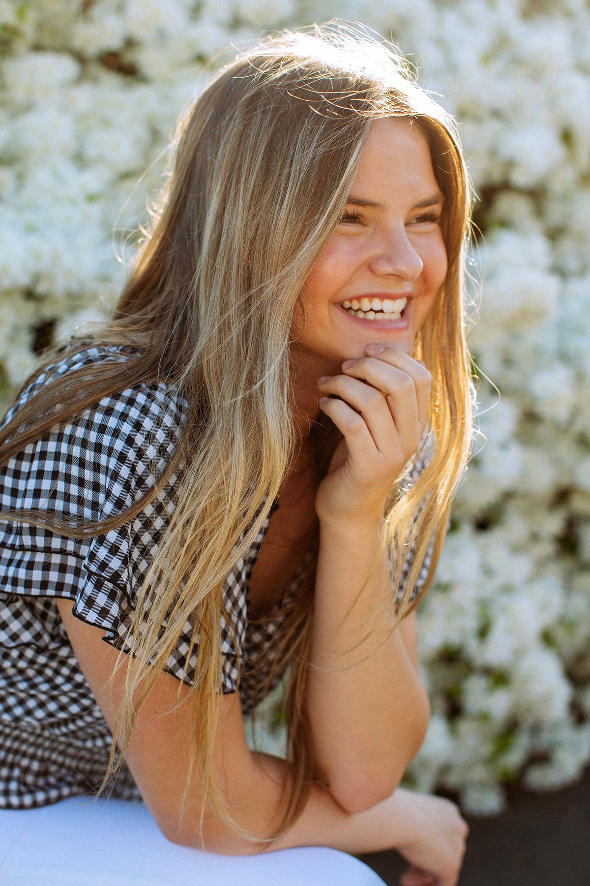 Smiling blonde teen girl in gingham top laughing outdoors near white flowering bushes in sunlight.