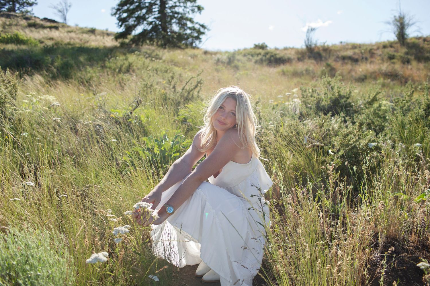 Blonde woman in white dress sitting in tall grass in a sunlit natural outdoor field setting.