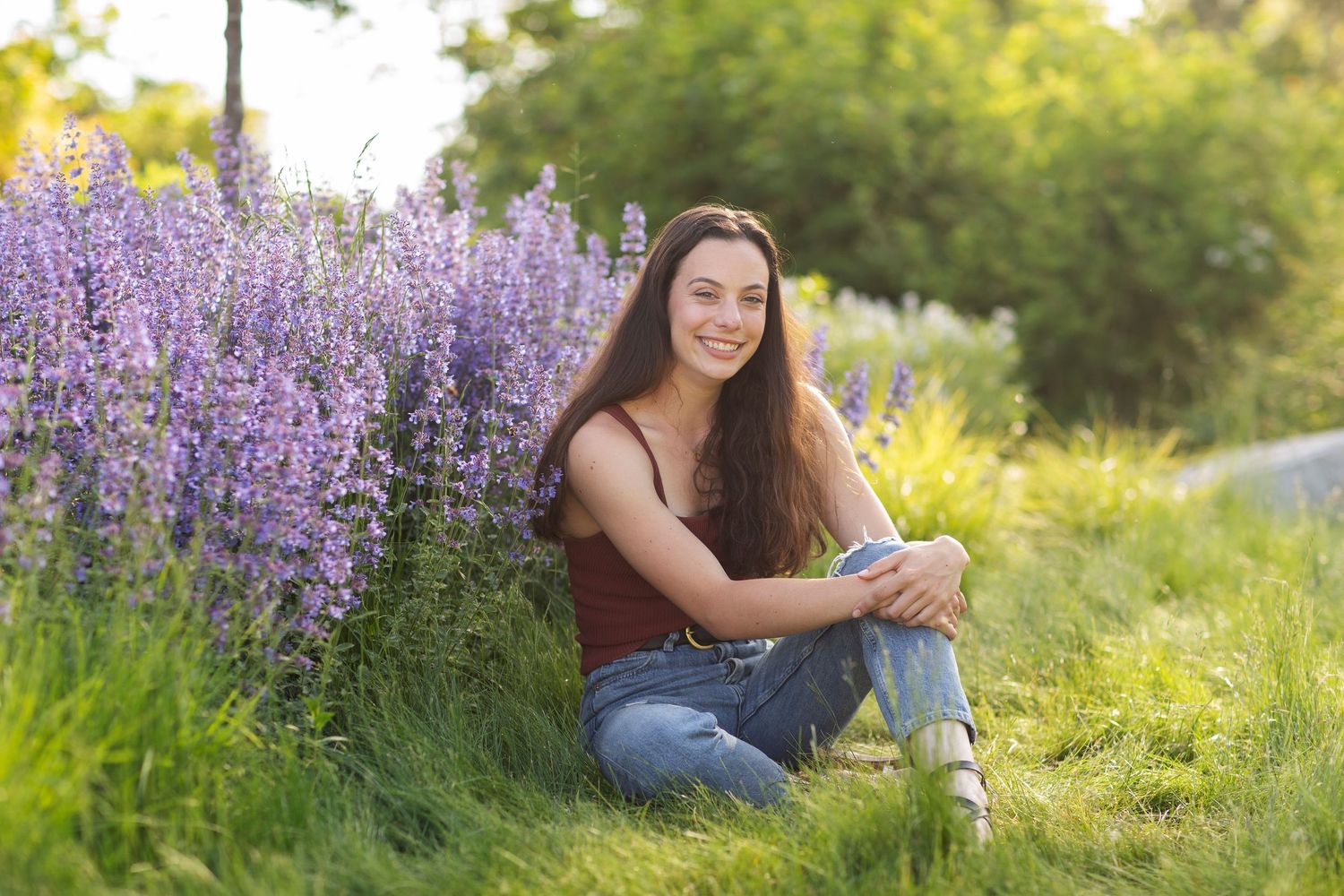 Young woman sitting on grass beside purple lavender flowers, smiling outdoors in a sunny garden setting.