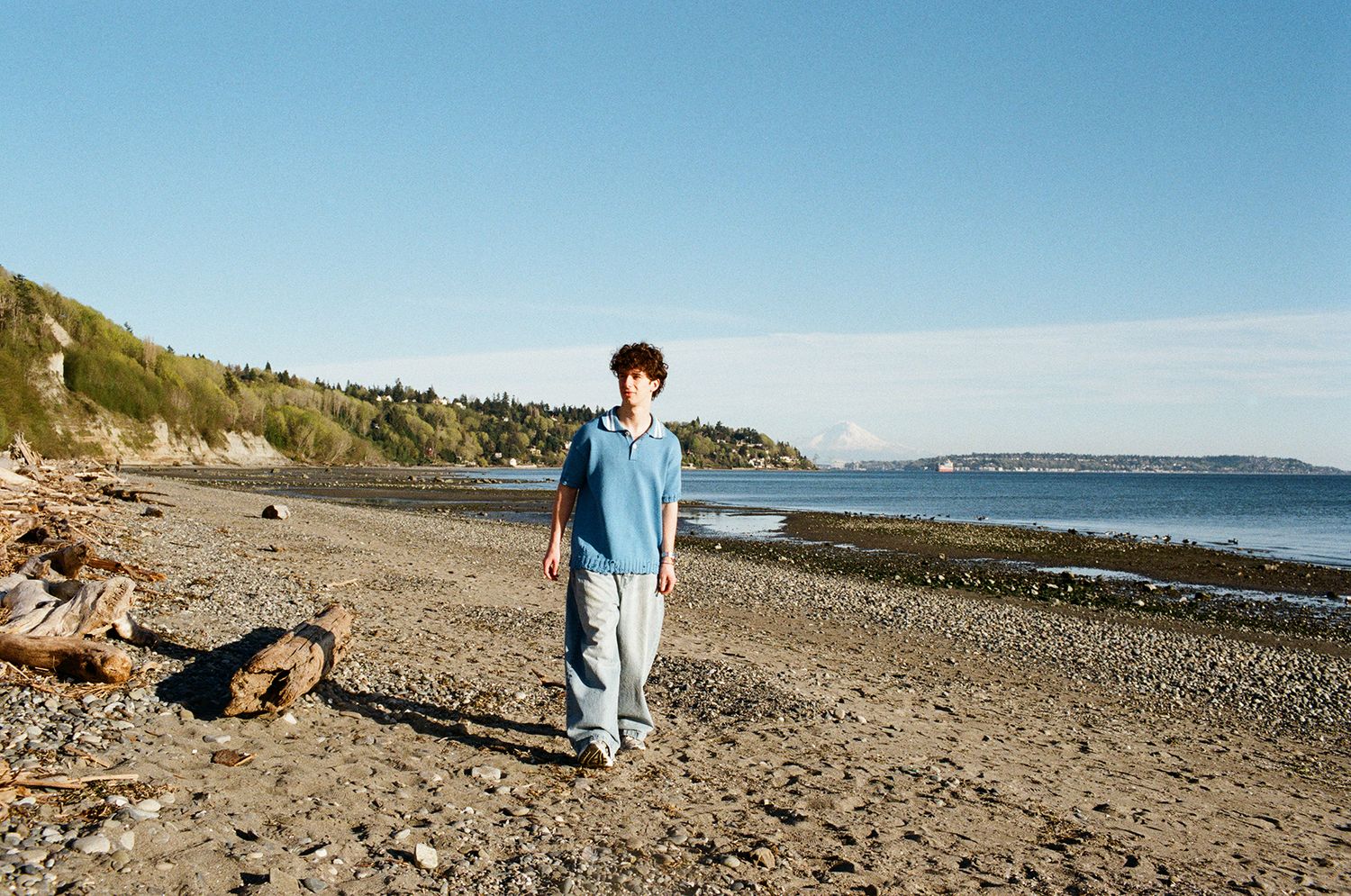 A young person stands alone on a rocky beach shoreline with calm water and green hills in the background.