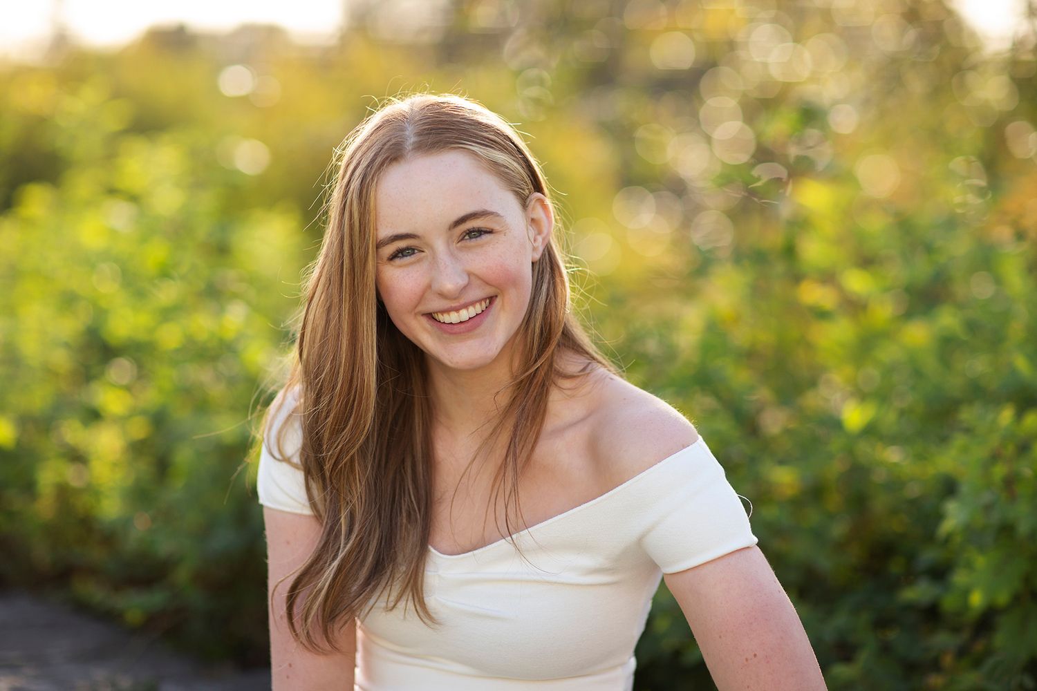 Smiling young woman with long hair wearing white off-shoulder top, posing outdoors with green foliage background.