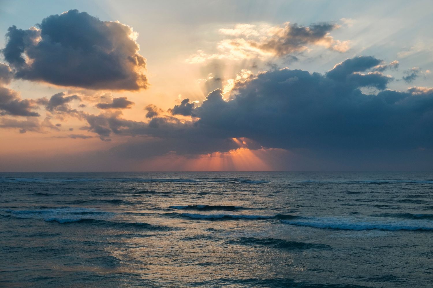 Stunning ocean sunset with dramatic clouds and golden sun rays reflecting on calm blue waves.