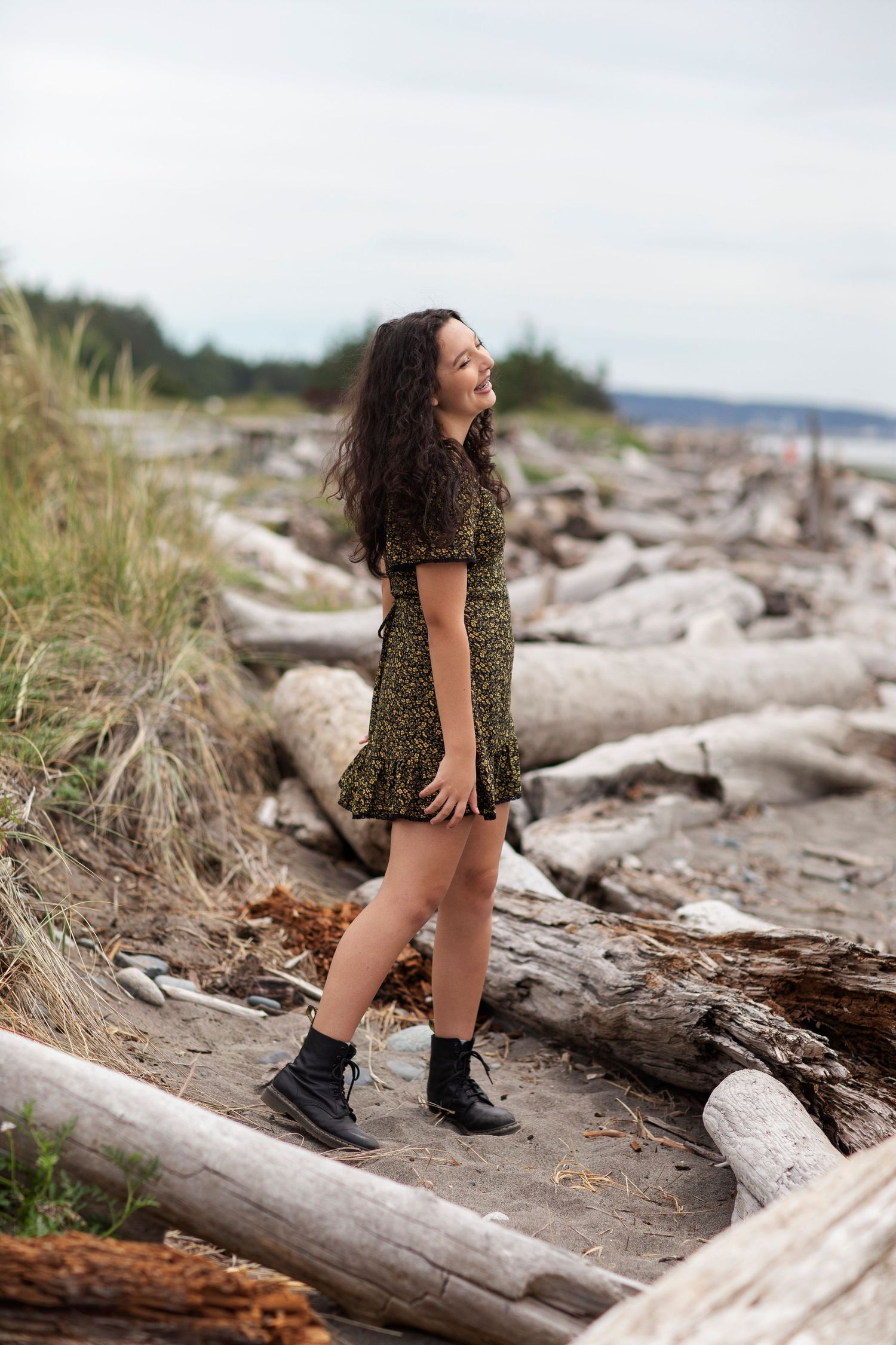 Young woman in green floral dress and black boots standing on driftwood at beach.