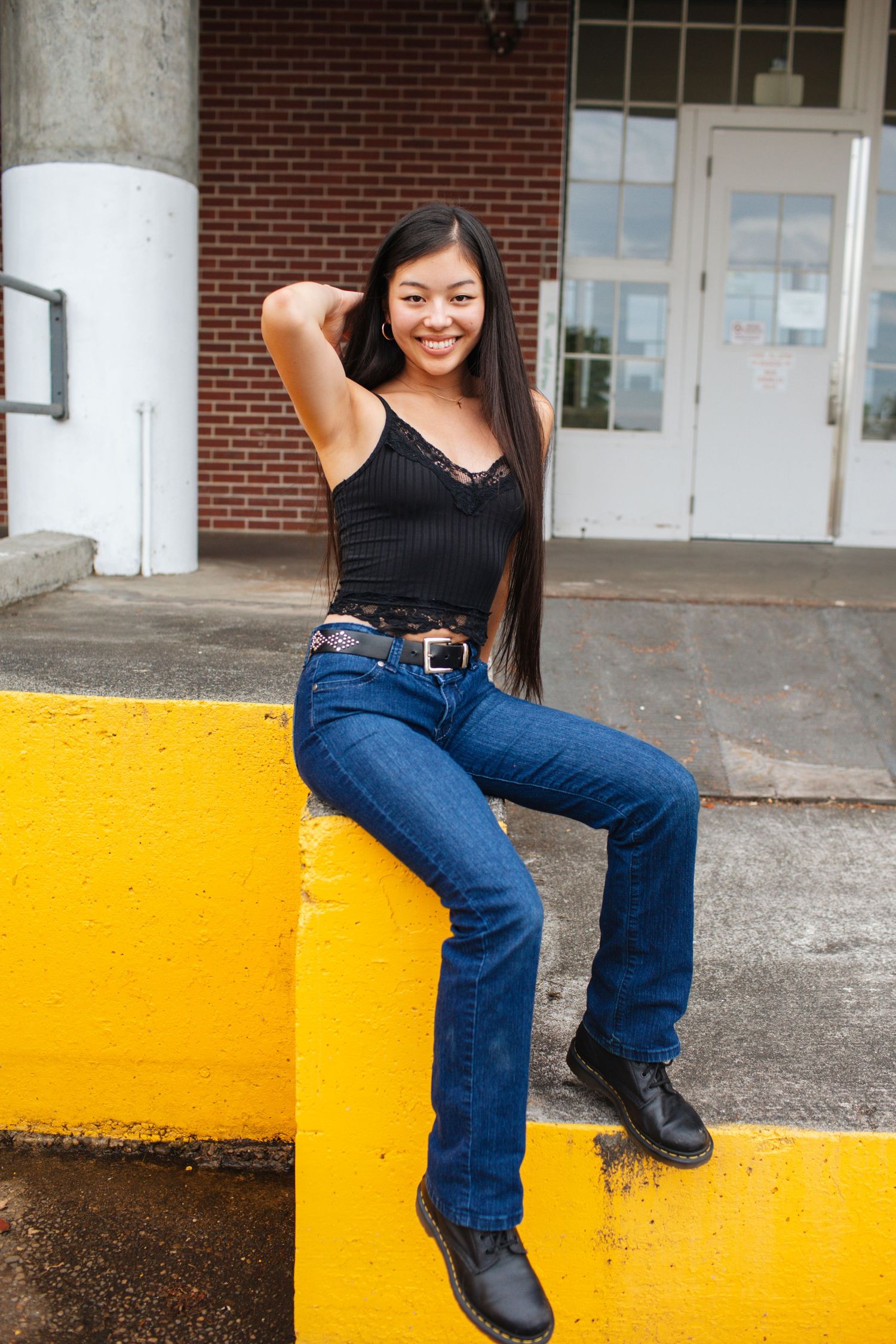 Smiling young woman with long dark hair sitting on yellow barrier outside brick building.