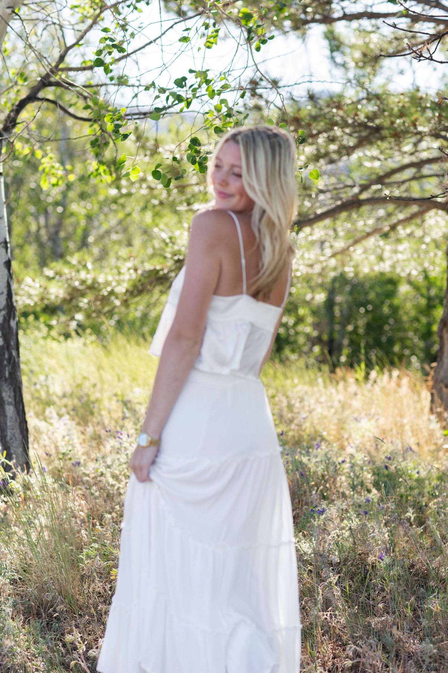 Blonde woman in white maxi dress standing among trees in a sunlit meadow, glancing over her shoulder.