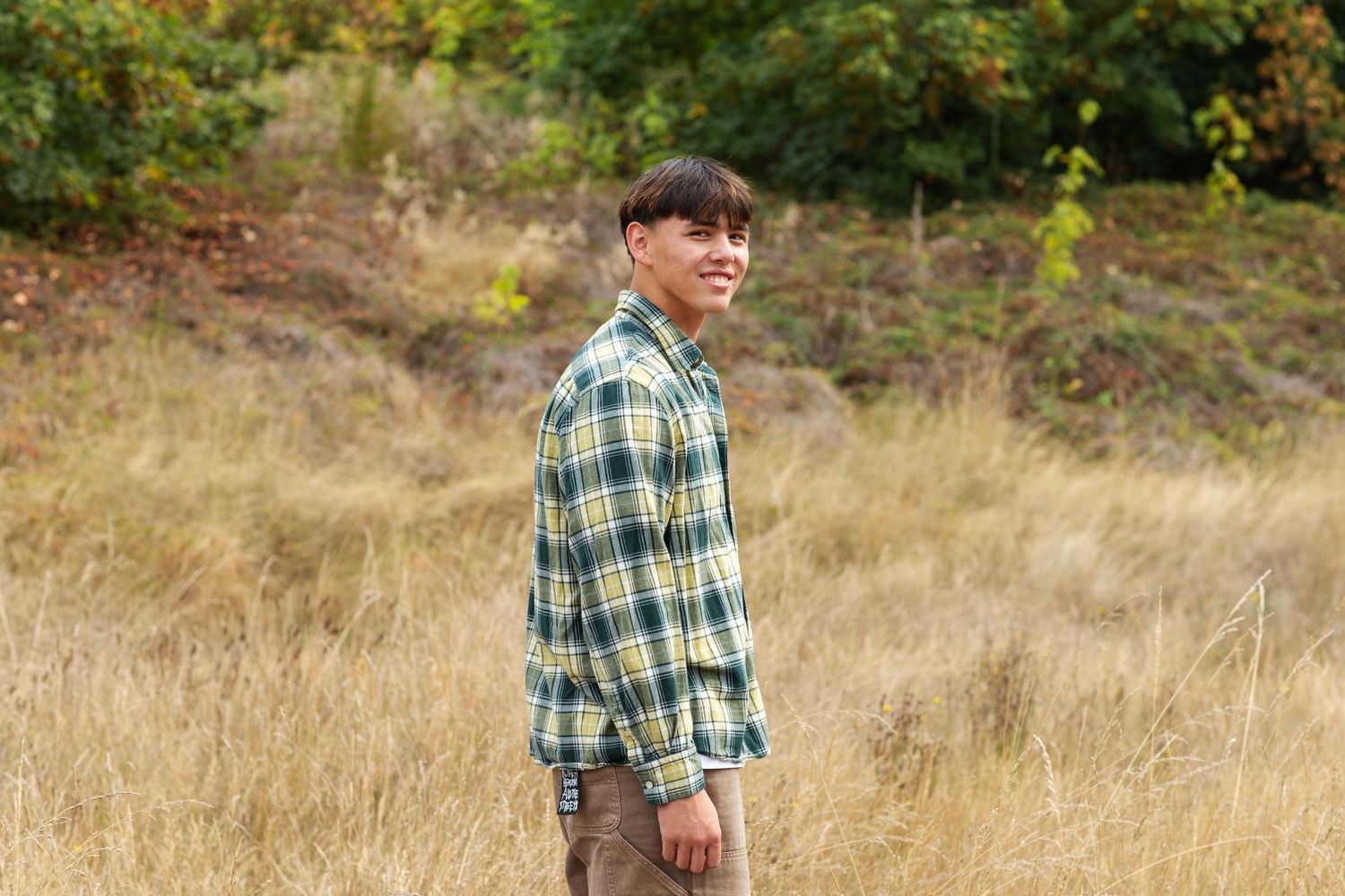 Young man in green plaid shirt standing sideways smiling in golden autumn field.