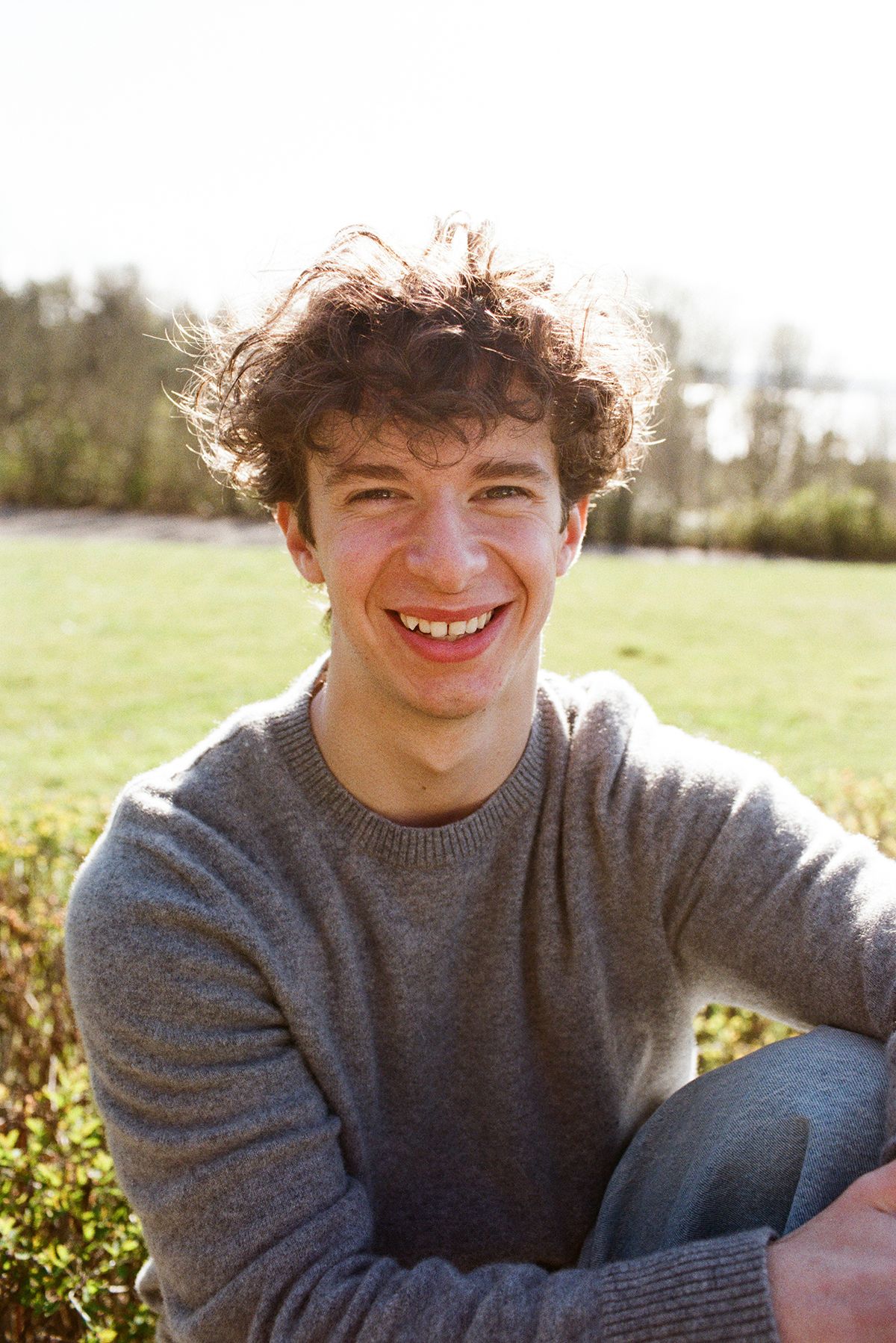 Smiling young man with curly hair wearing a gray sweater sitting outdoors on a sunny day.