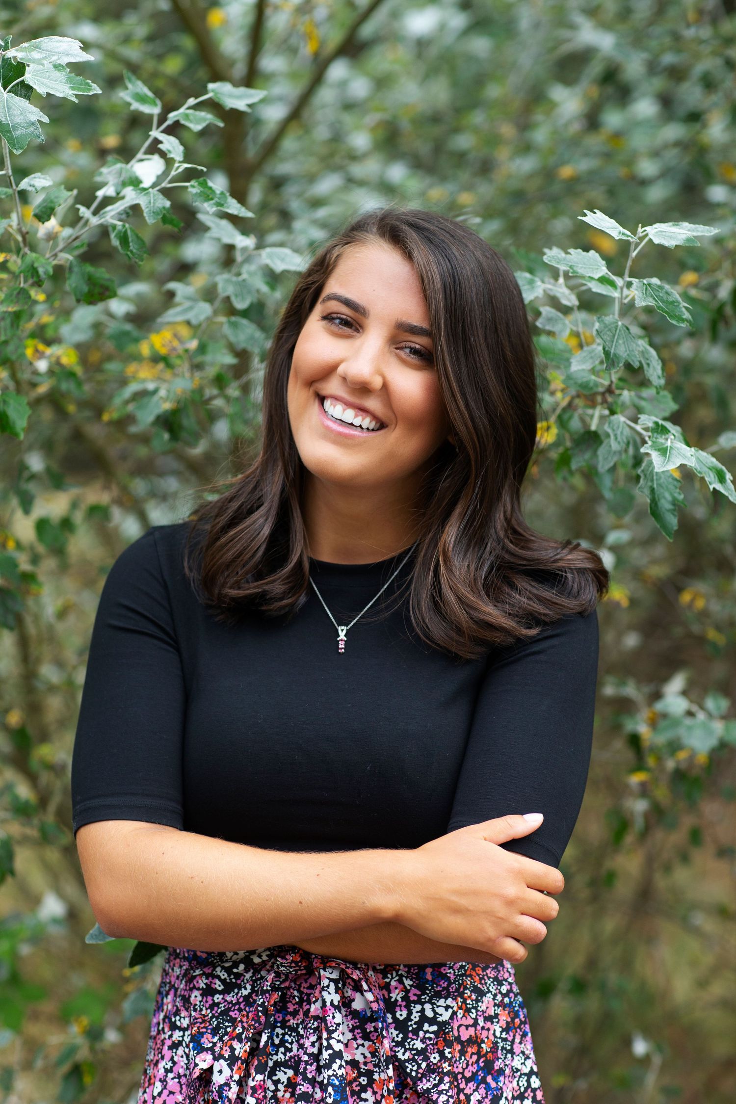 Smiling young woman in black top and floral skirt posing with arms crossed in front of green foliage.