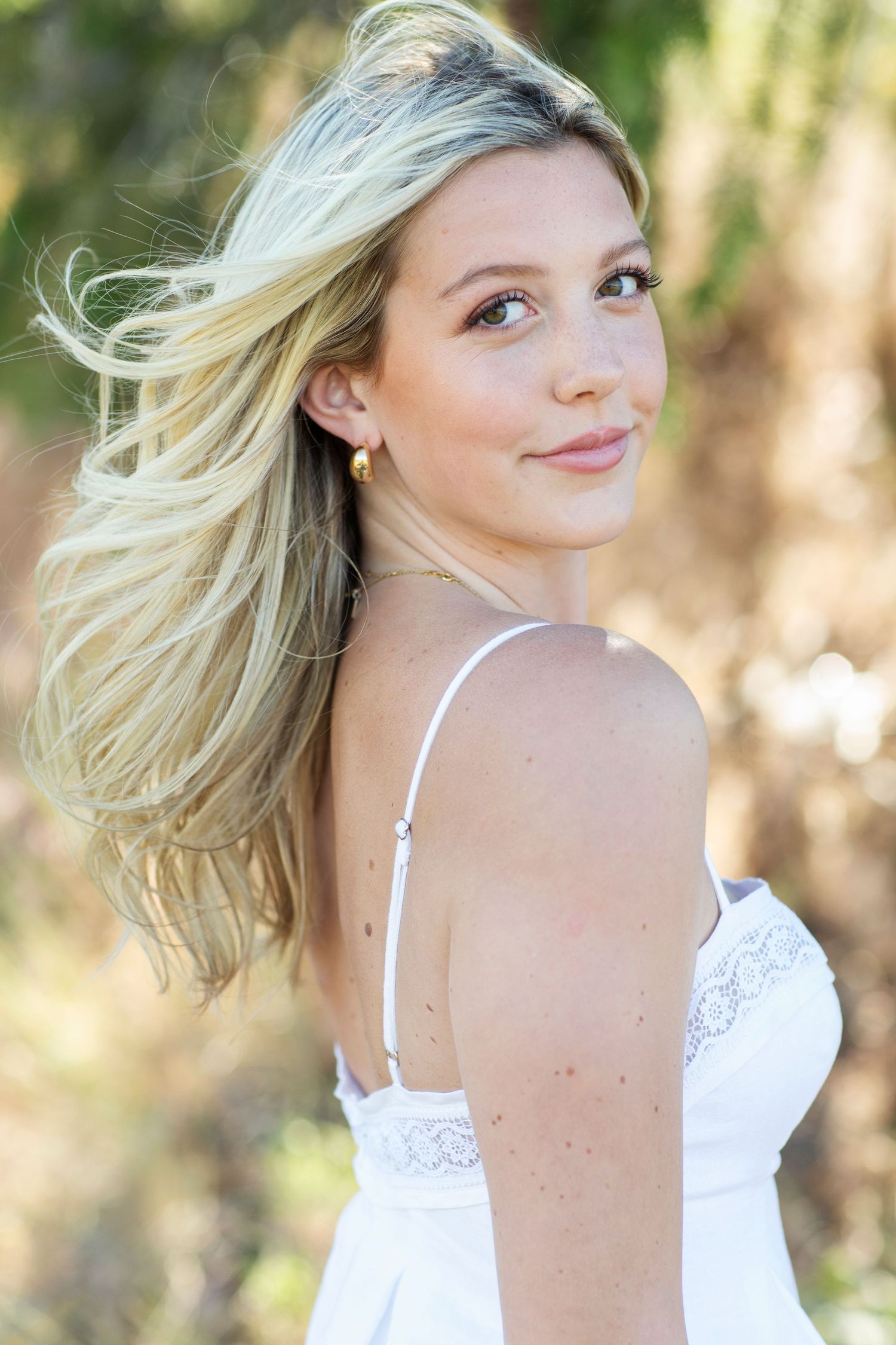 Close-up of a blonde woman in a white lace dress glancing over her shoulder with a soft smile outdoors.