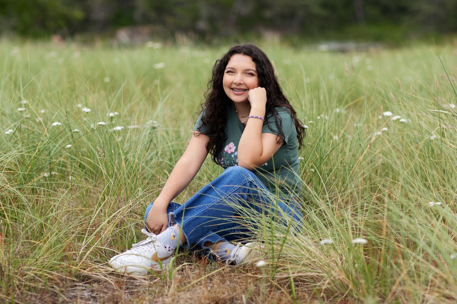 Smiling teen girl sitting in tall grass field wearing green tee and jeans.