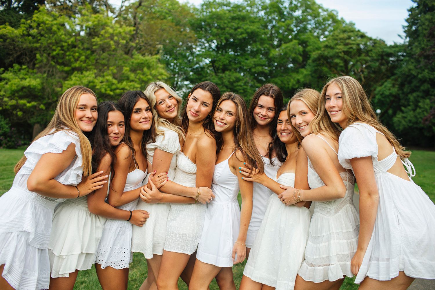 Group of young women in matching white dresses smiling and posing together outdoors on a sunny day.