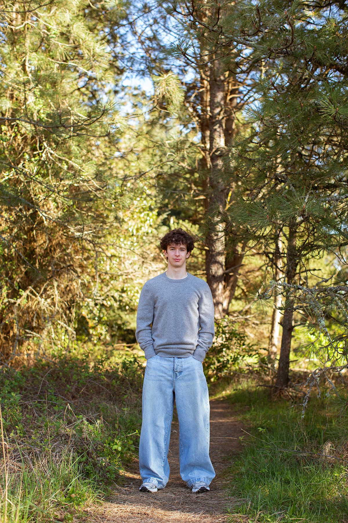 Young man in gray sweatshirt and wide-leg jeans standing on forest path surrounded by pine trees.