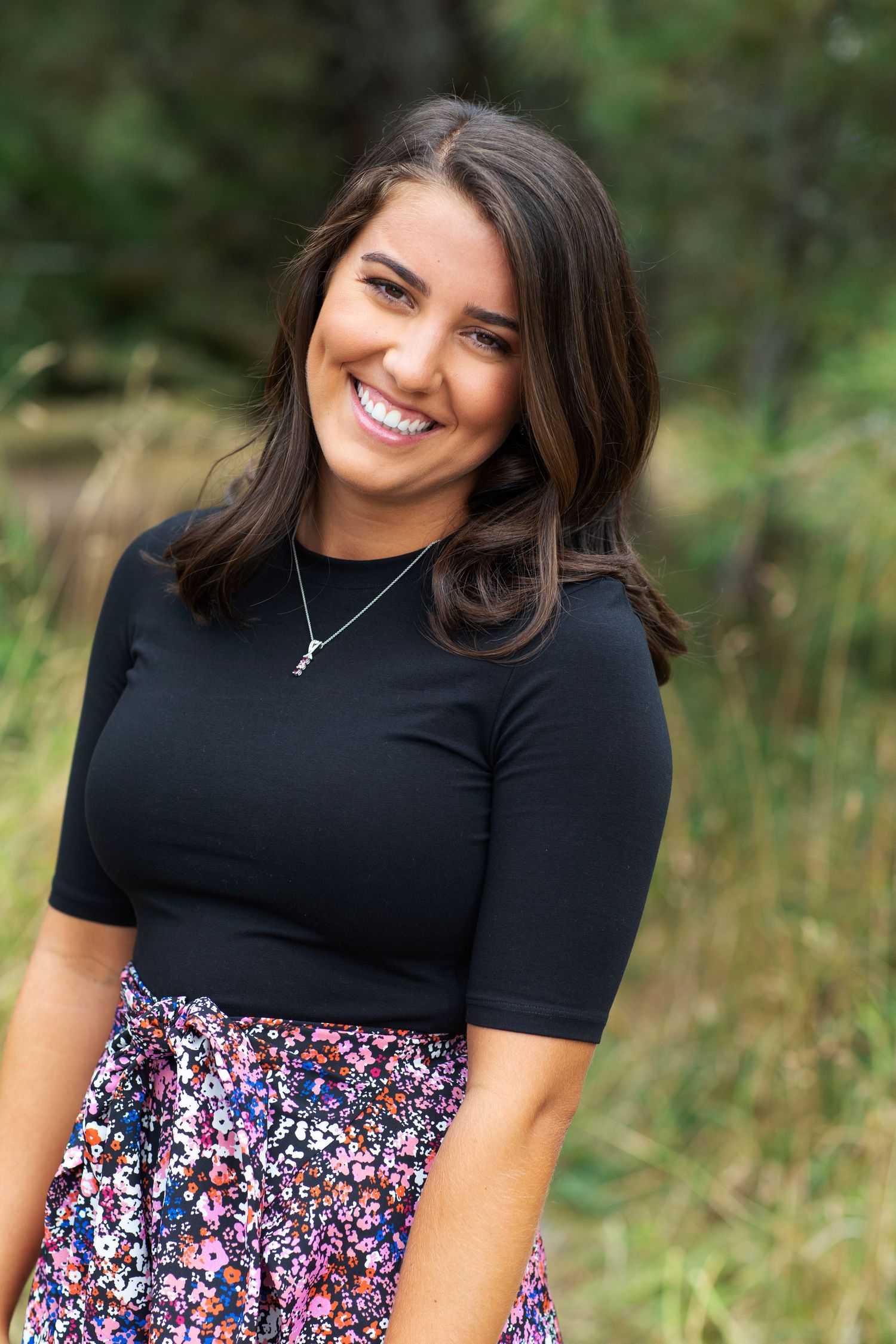 Smiling brunette woman in black top and floral skirt outdoors.