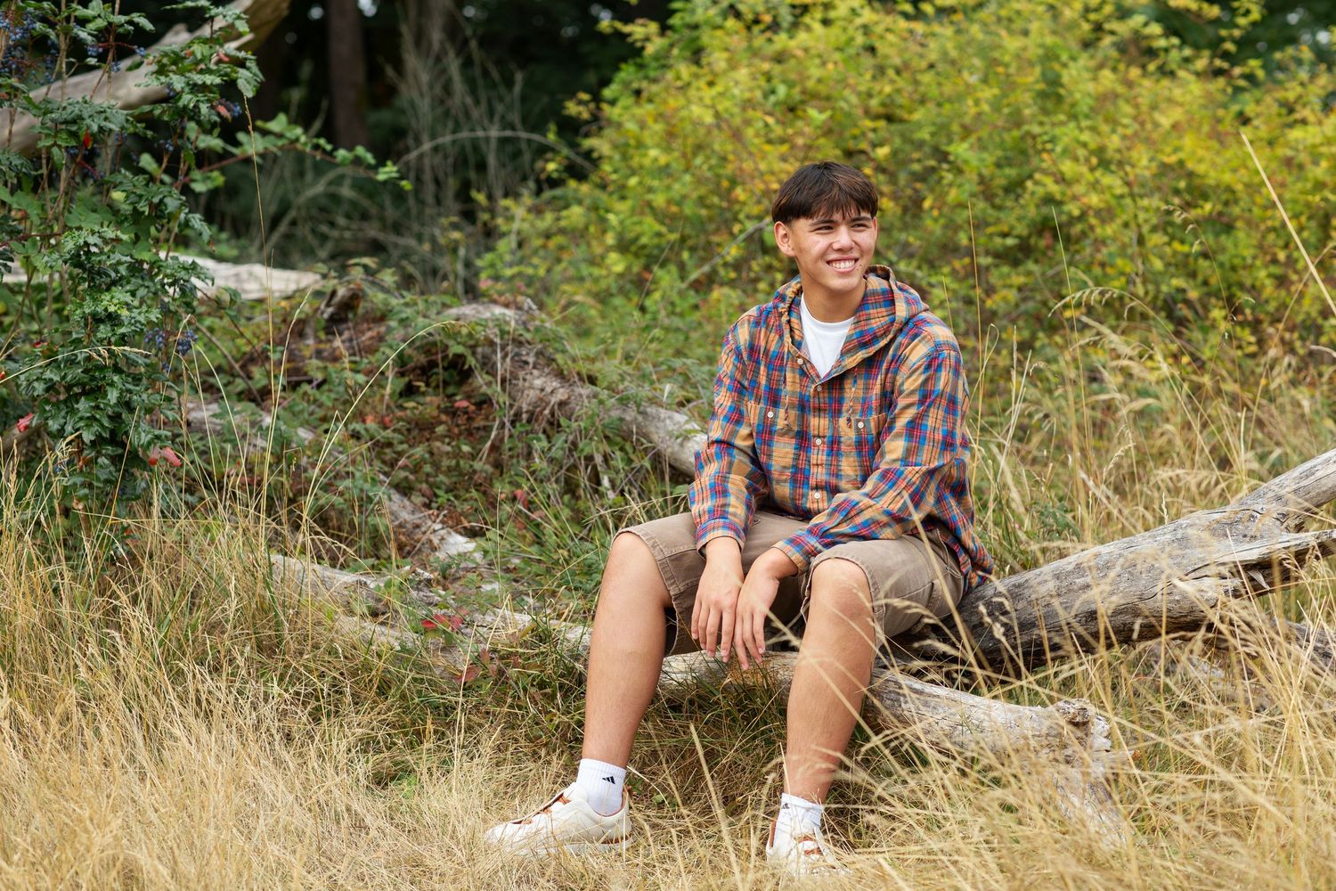 Teen boy in plaid flannel shirt sitting on log in golden grassy field outdoors.