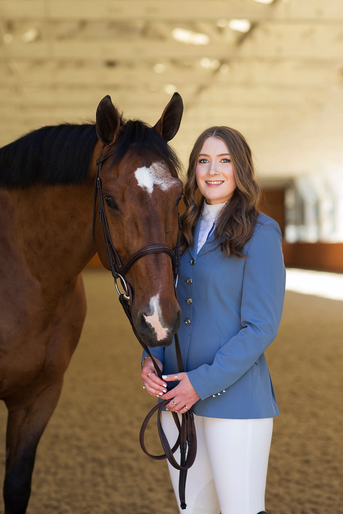 A smiling young woman in a blue equestrian jacket stands beside a brown horse with a white blaze in an indoor riding arena.