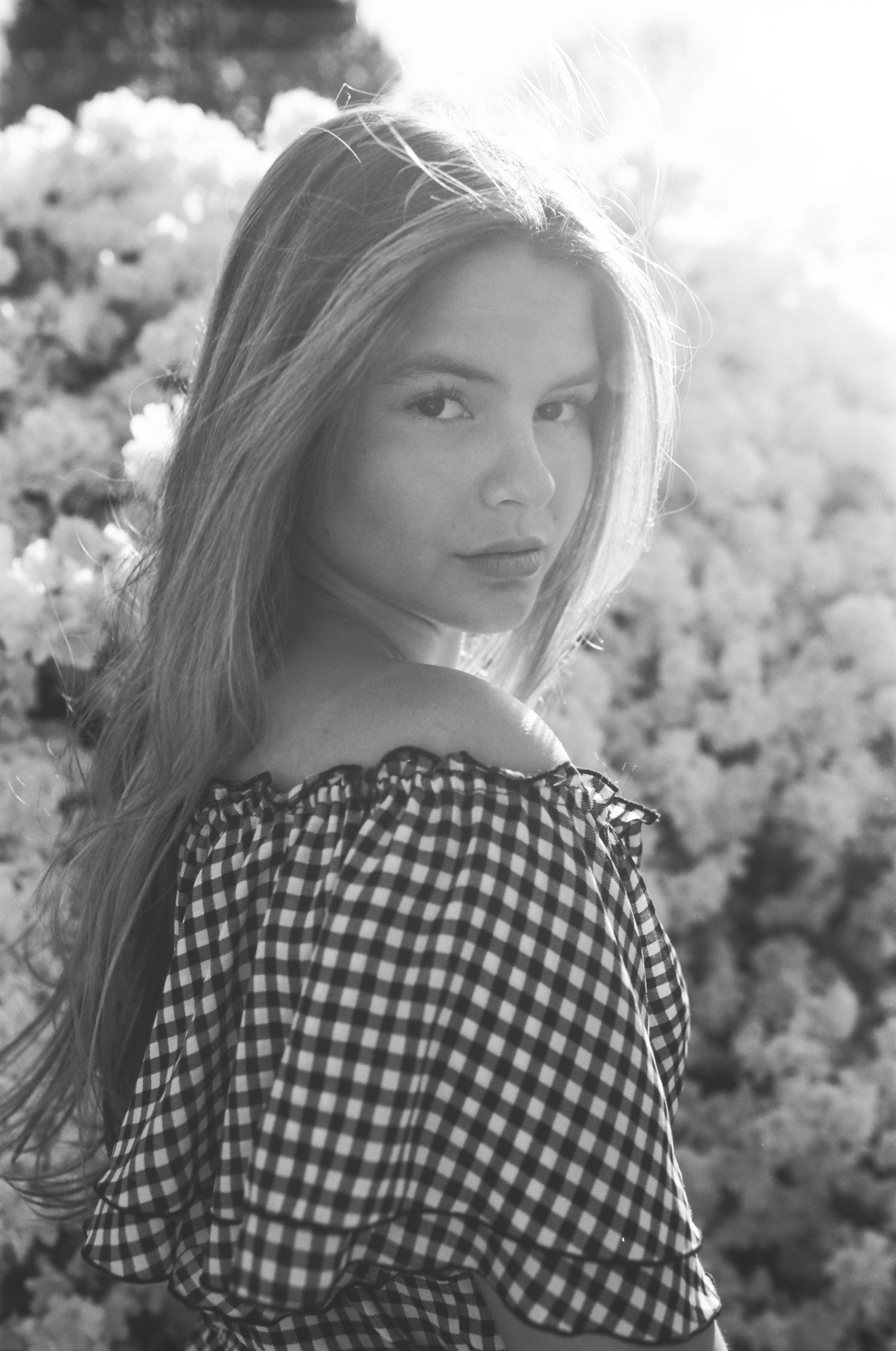 Black and white portrait of young woman in gingham off-shoulder top among blooming flowers.