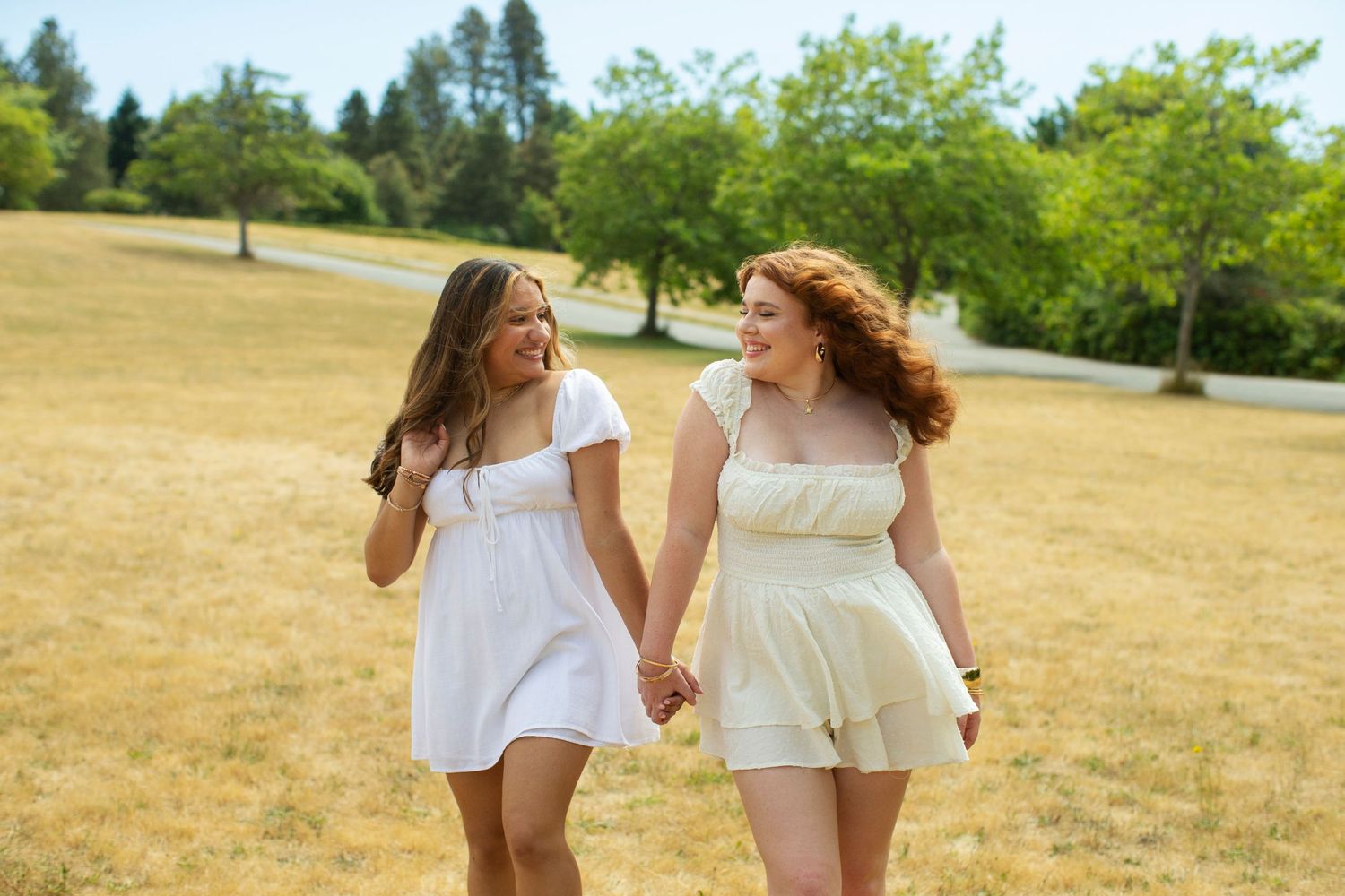 Two young women in white dresses holding hands walking in sunny field.